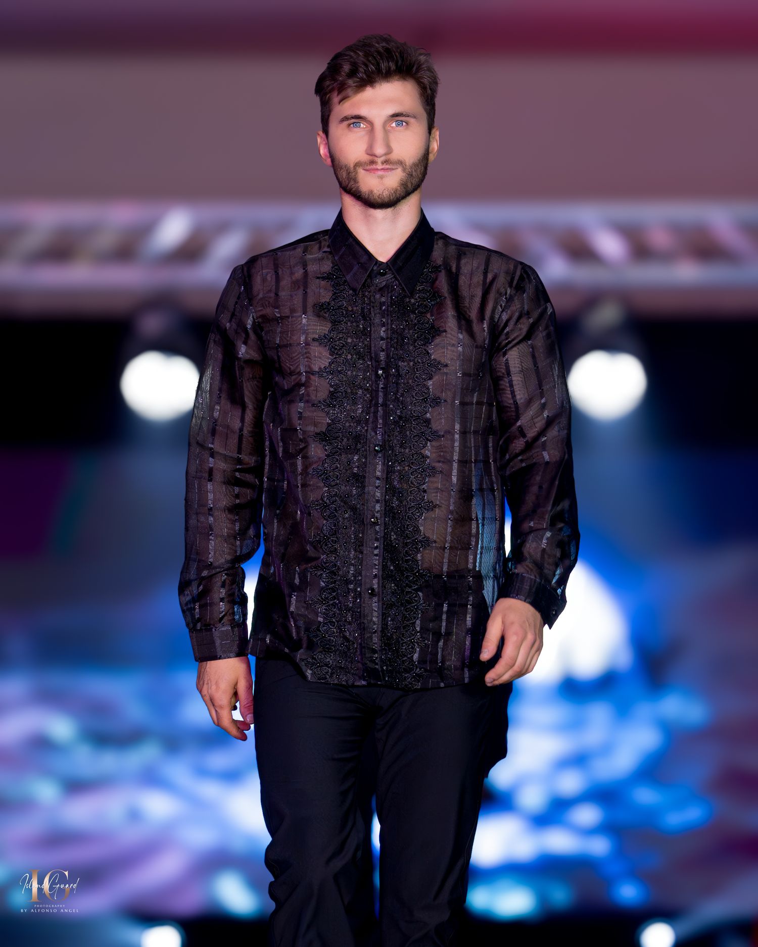 Male model wearing sheer black button-down shirt and dark pants on illuminated runway.