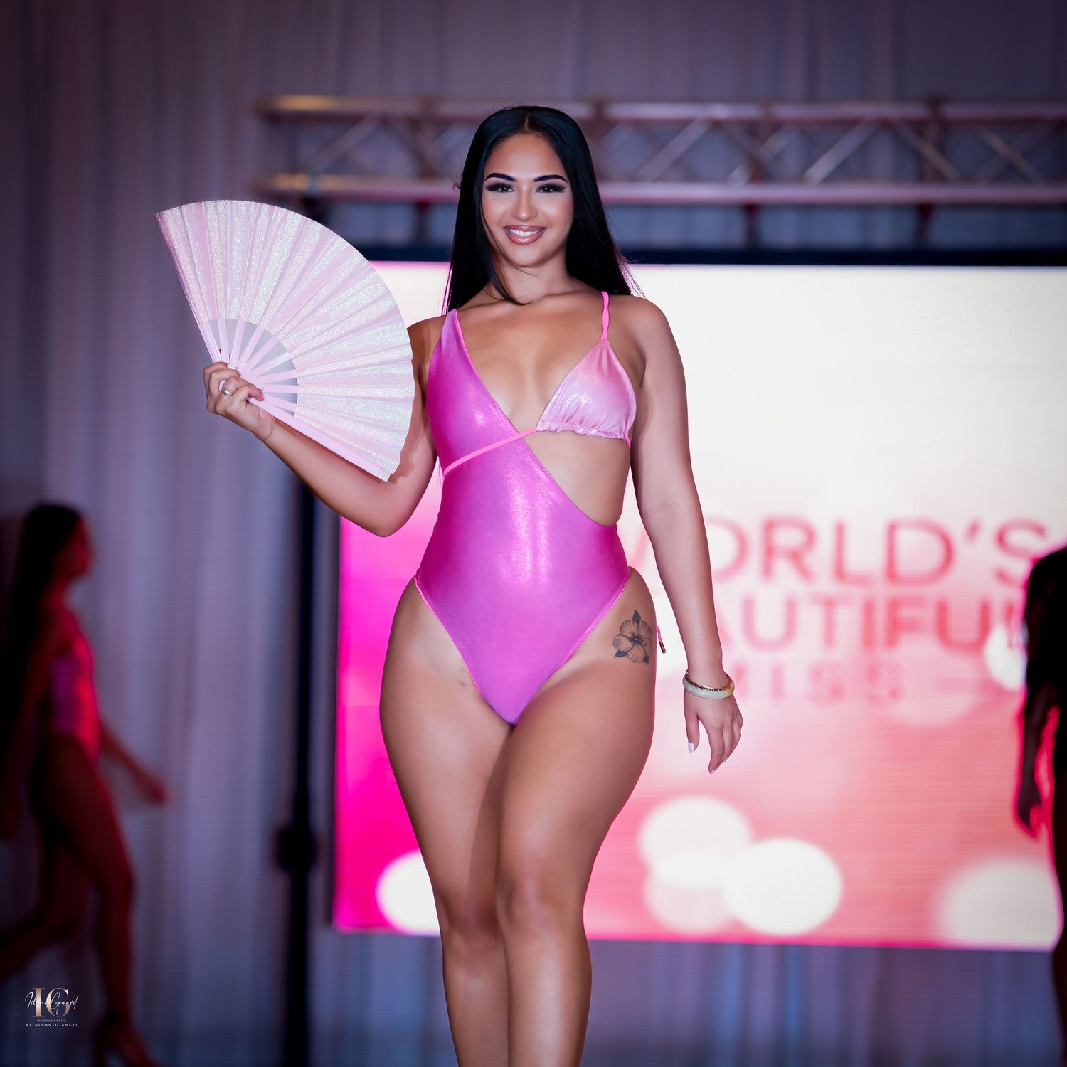 A model walks the runway in a bright pink swimsuit while holding a matching fan at a fashion show.