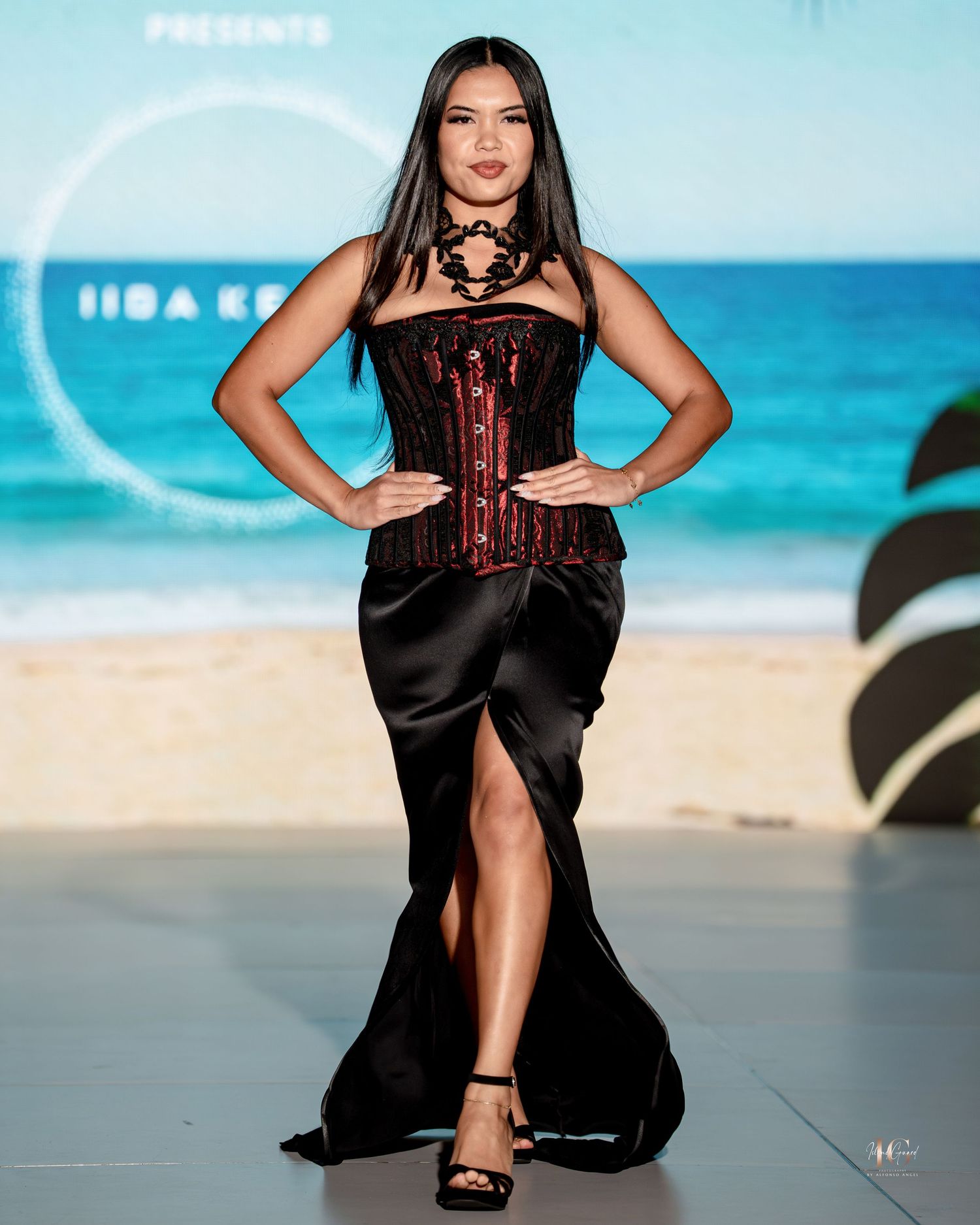Model walks runway in black and burgundy satin evening gown with high slit at beachside fashion show.