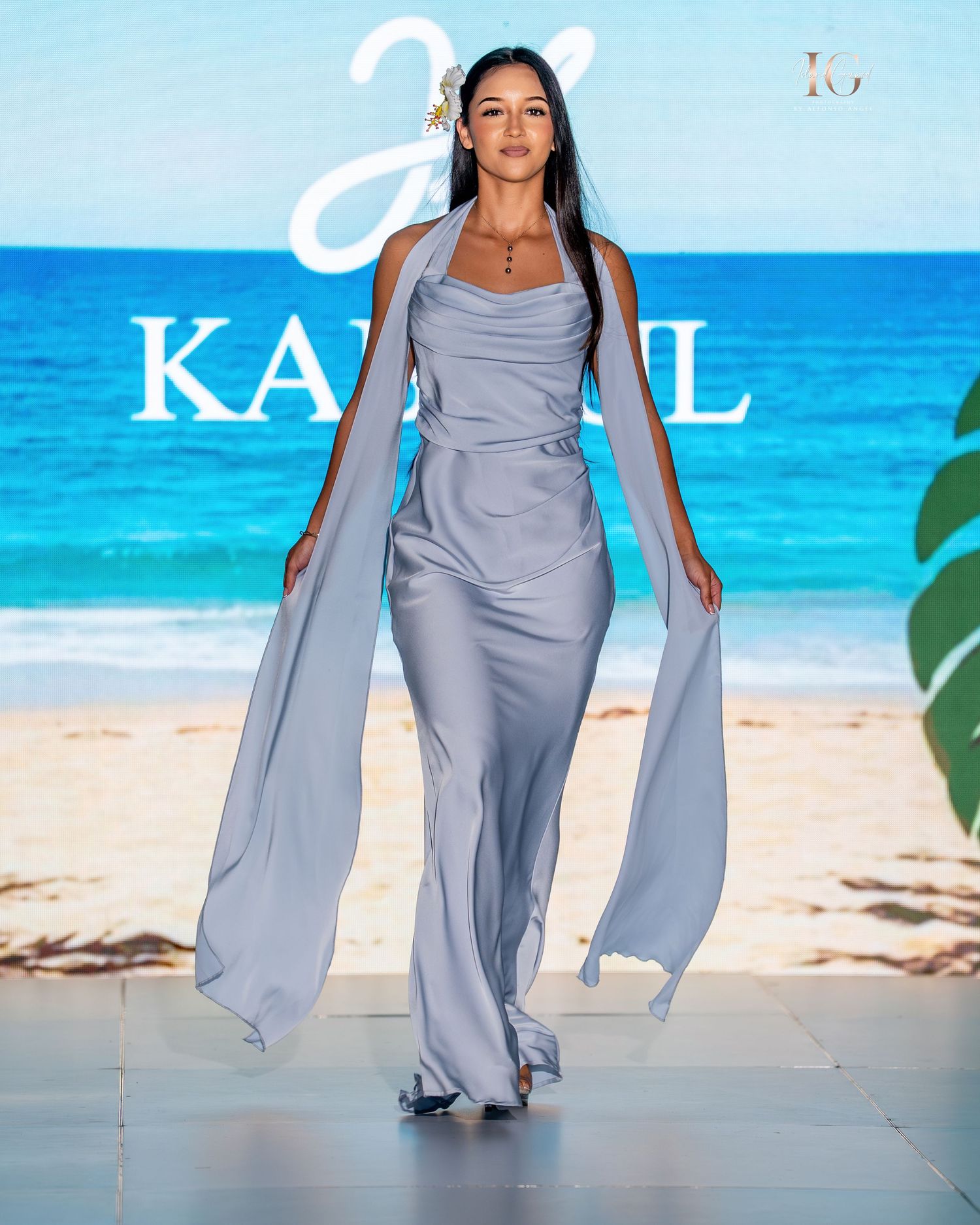 Model walks runway in flowing powder blue evening gown with cape sleeves on tropical beach backdrop.