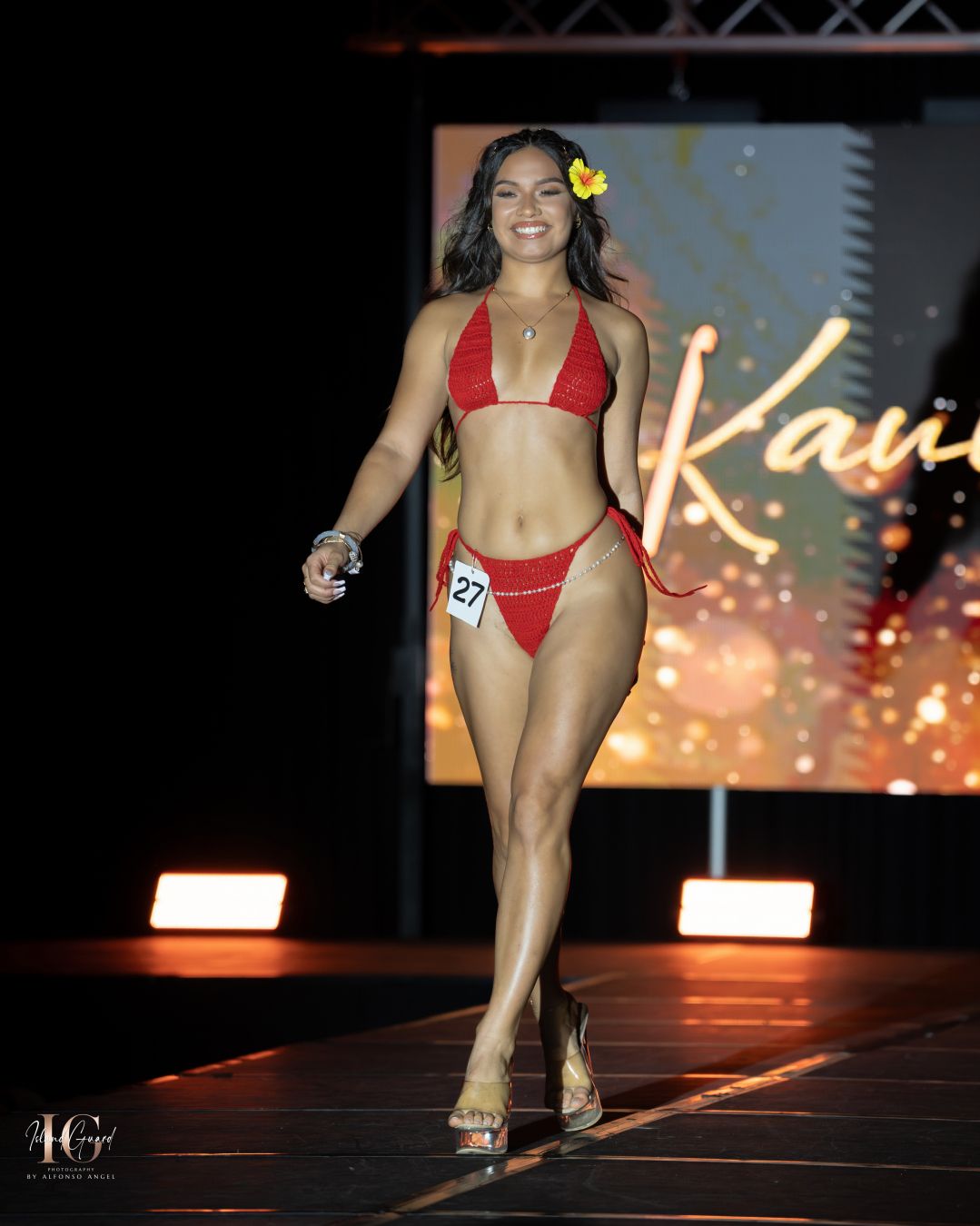 Model in red bikini walks runway with yellow flower accessory during fashion show event.