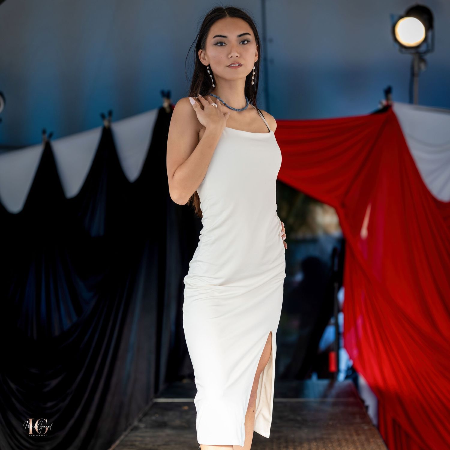 Model on runway wearing short white dress with red backdrop draping.