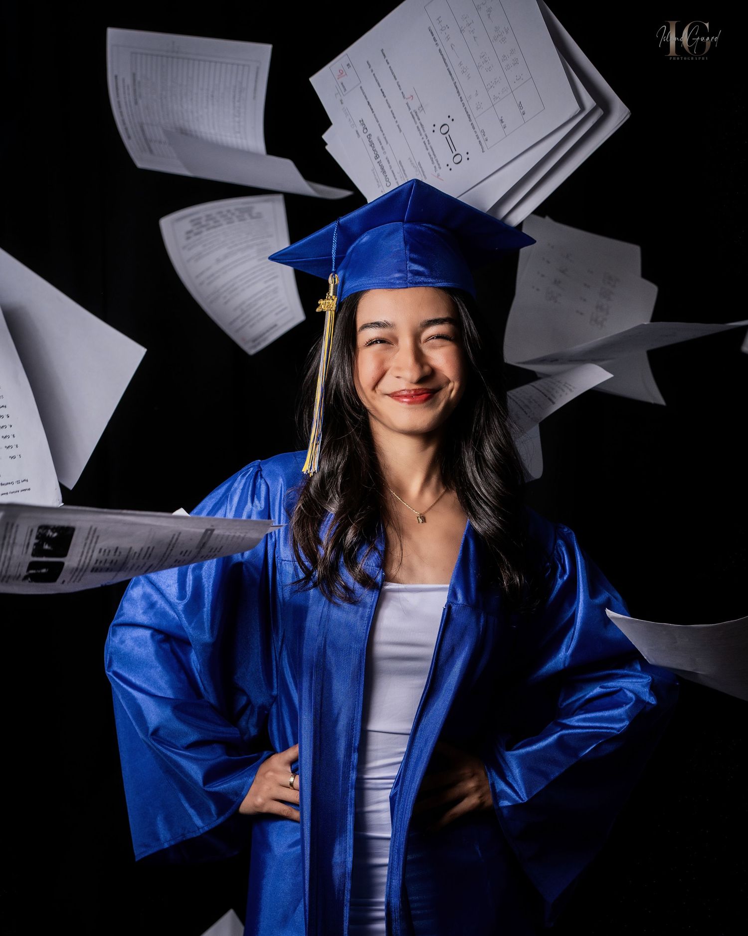 Graduate wearing blue cap and gown holds up academic papers while smiling in a dark studio setting with dramatic lighting.