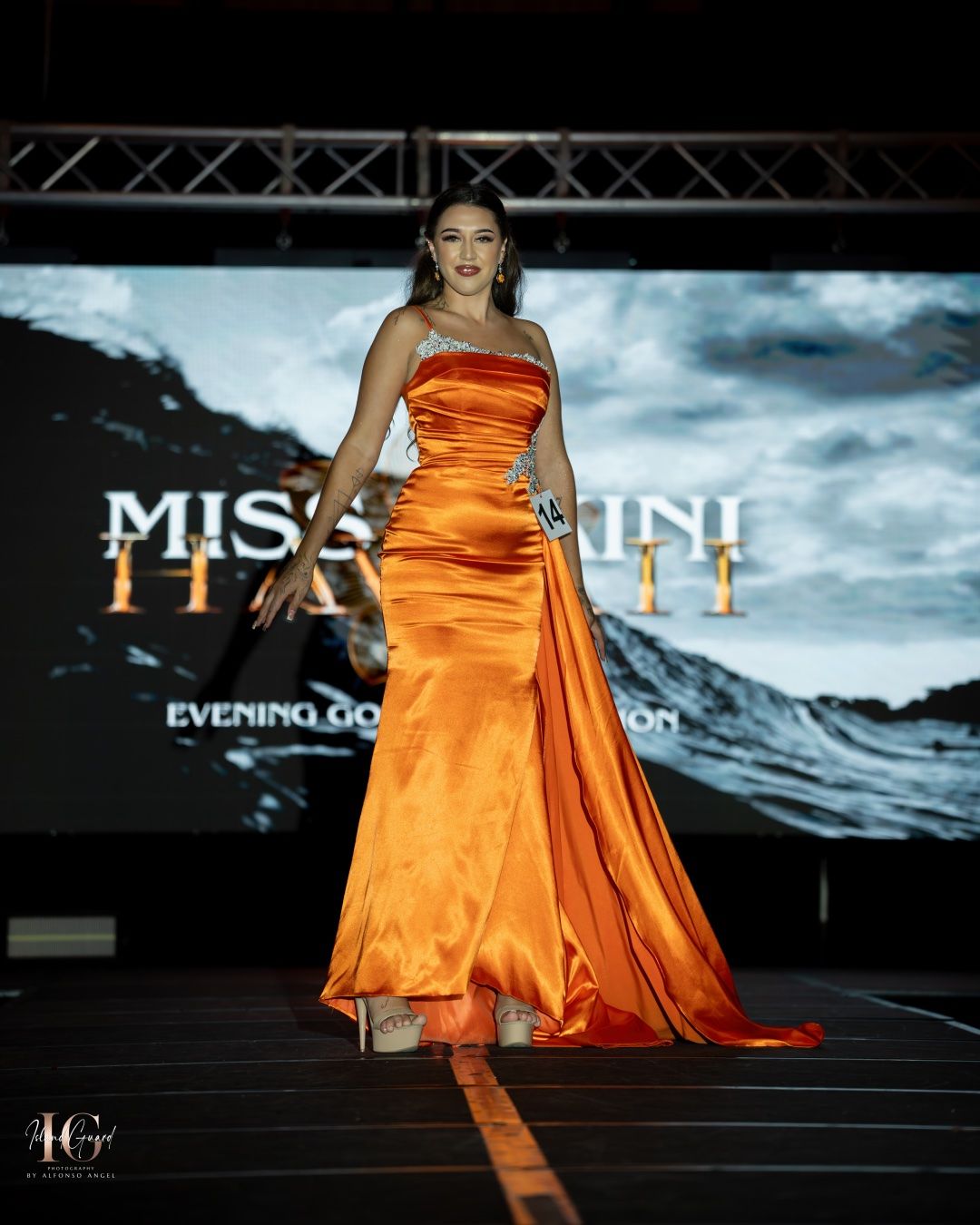 Models walk the runway in vibrant orange satin evening gowns during a glamorous fashion show presentation.