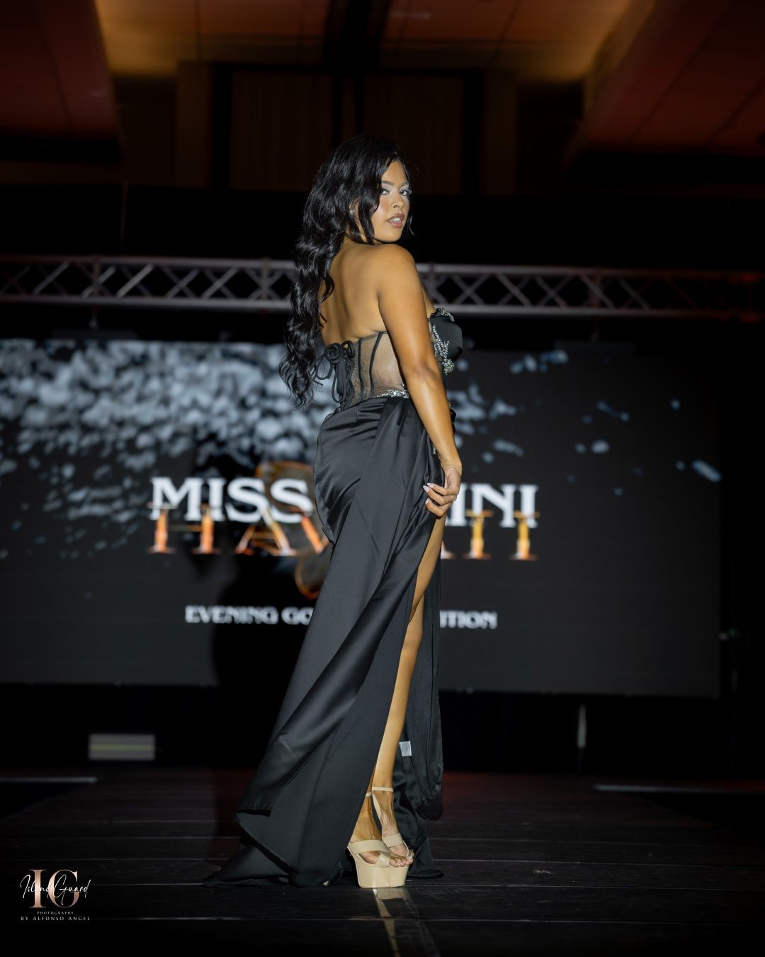 A model in a sleek black evening gown poses on stage at a pageant event with dramatic lighting and signage.