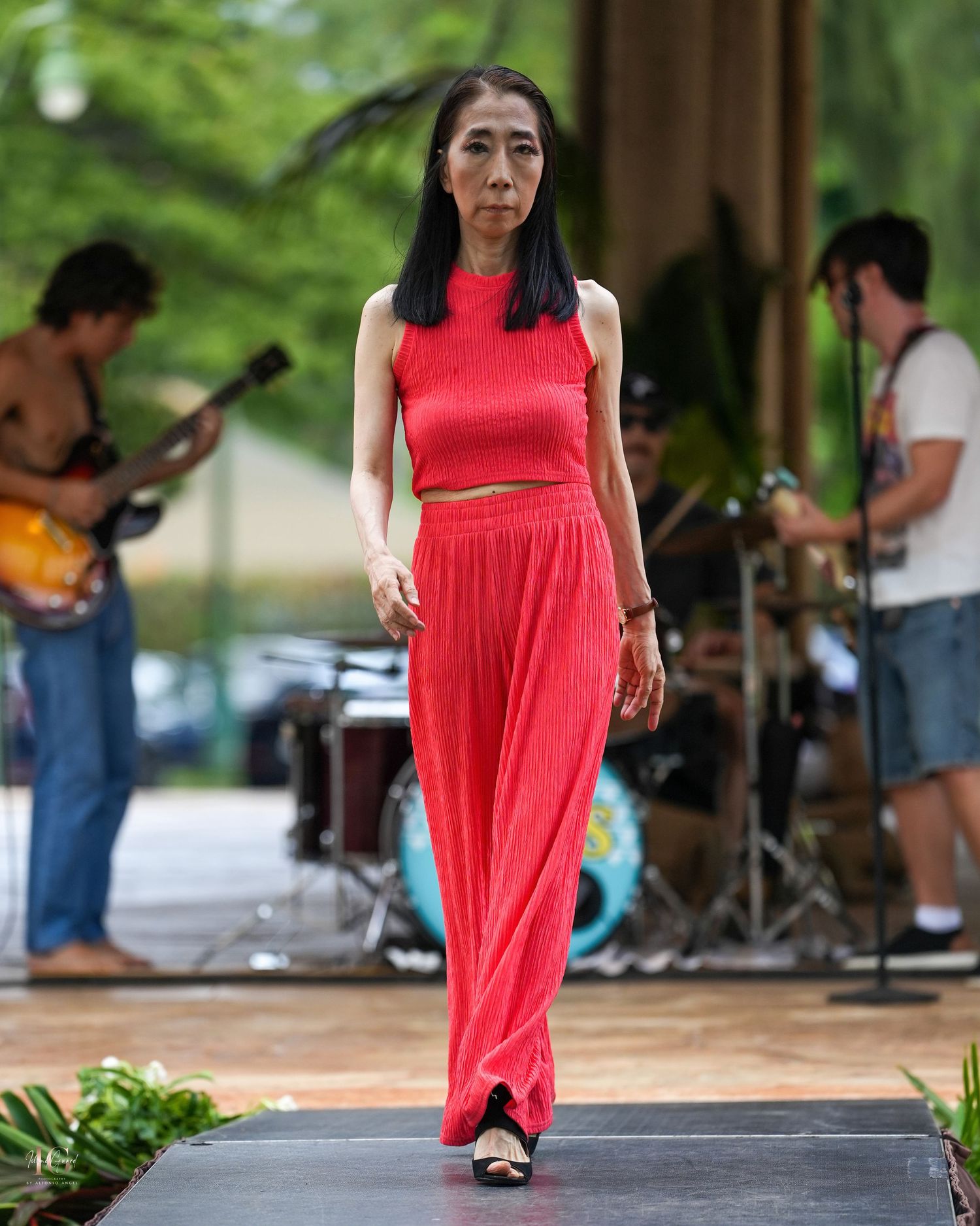 A person in a flowing red dress performs an expressive dance movement outdoors near trees and other performers.