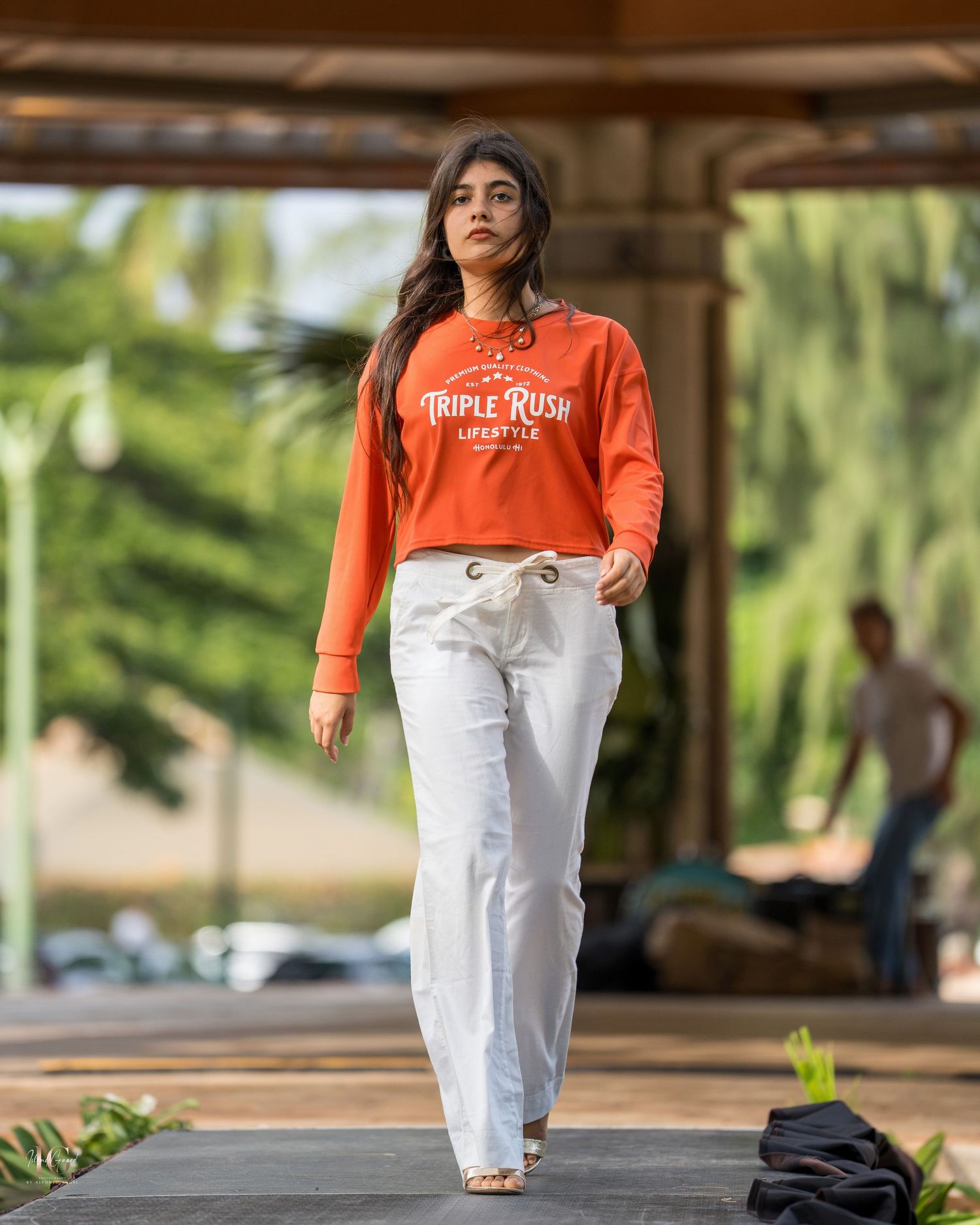 Someone wearing an orange sweatshirt and white jeans poses casually in an outdoor setting with trees and buildings.