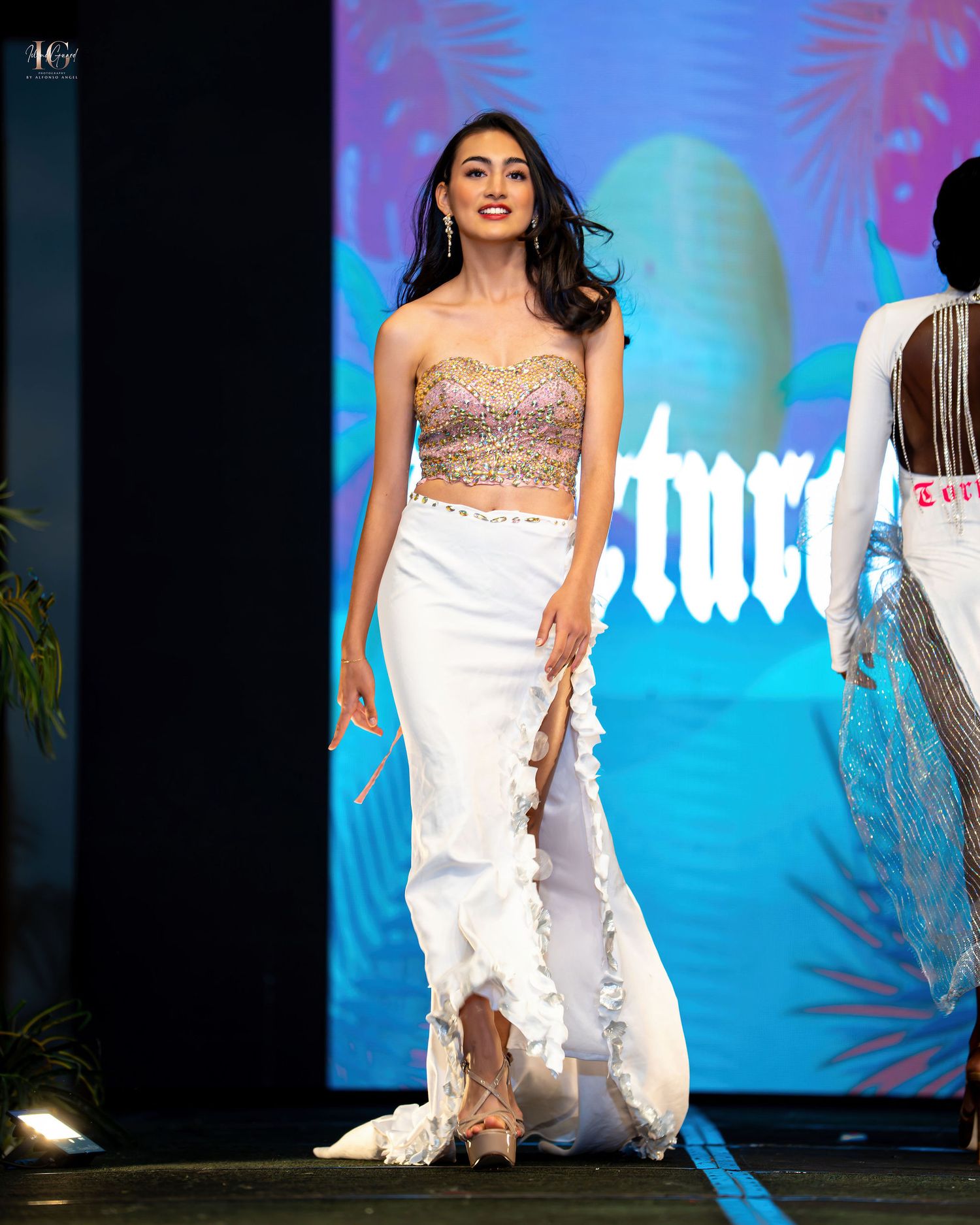 Fashion model walks the runway in a flowing white dress with embellished details against a vibrant blue backdrop.