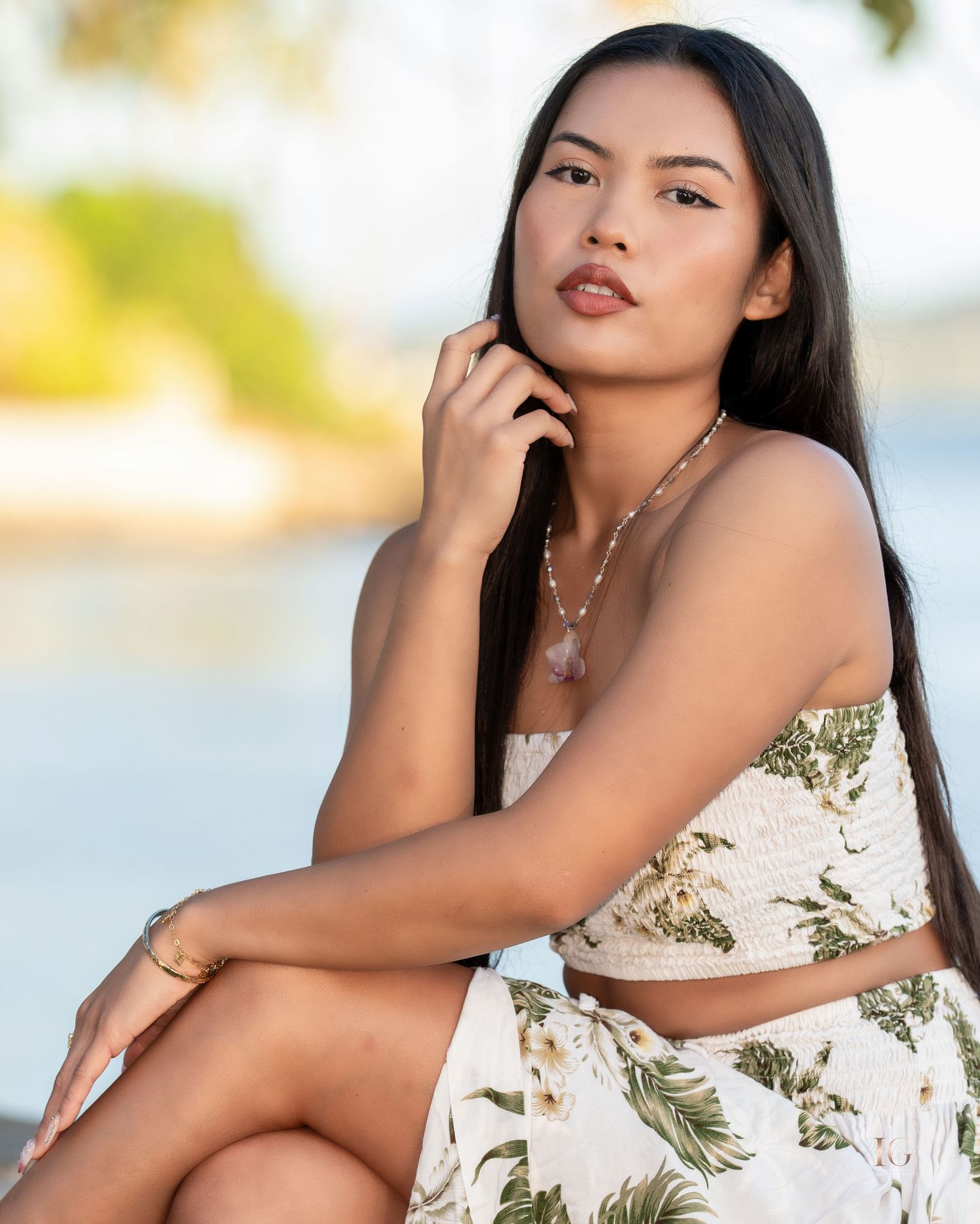 A person in a floral print dress poses outdoors in a tropical setting with palm trees in the background.