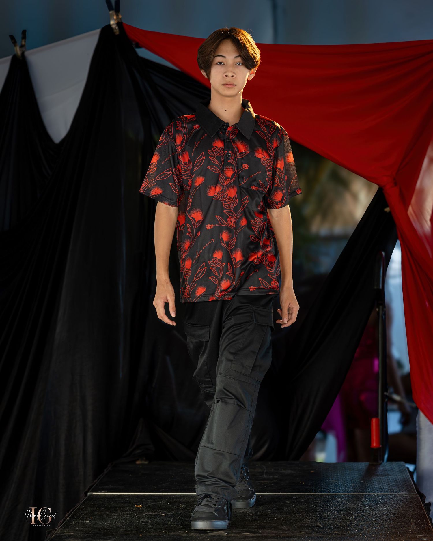 A runway model wearing a black and red patterned t-shirt with dark pants on stage.
