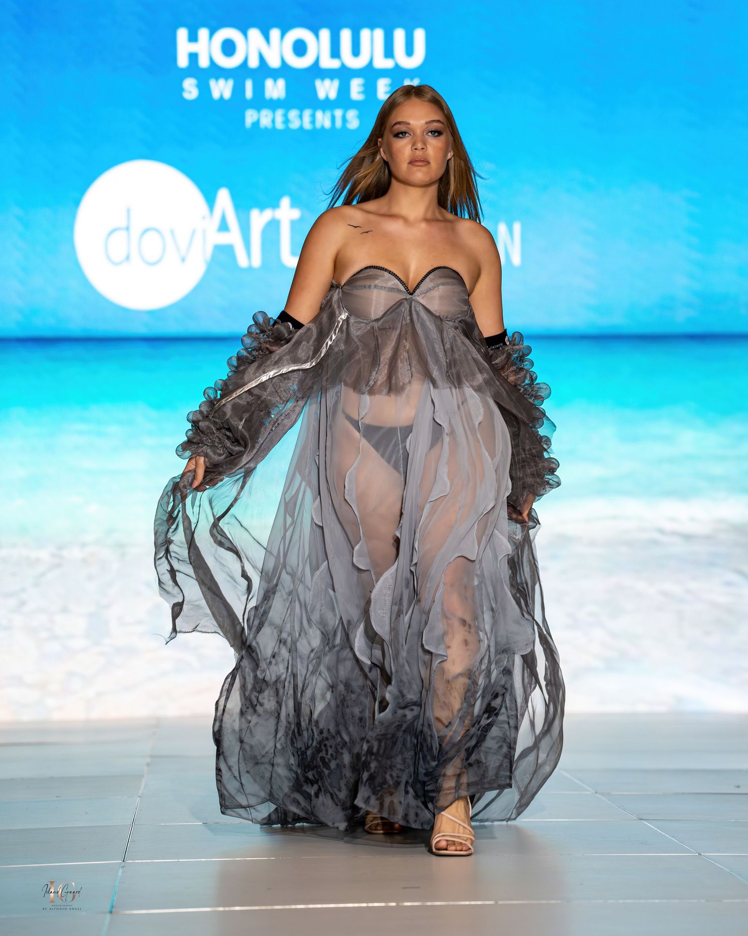 Fashion model in sheer gray ruffled evening gown walks runway against beach backdrop at Honolulu Swim Week.