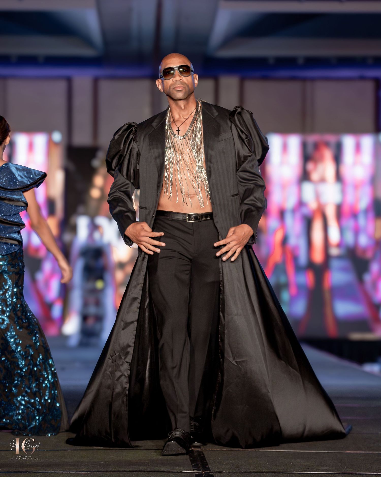 Dramatic black satin coat and sunglasses create bold silhouette on illuminated fashion runway.