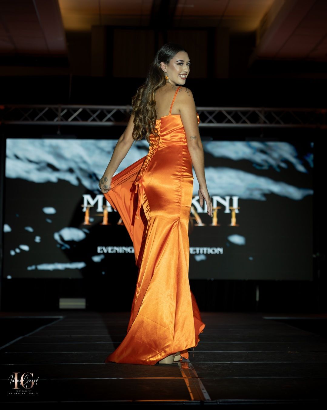 Model on runway showcasing orange satin dress with flared skirt in elegant fashion show presentation.