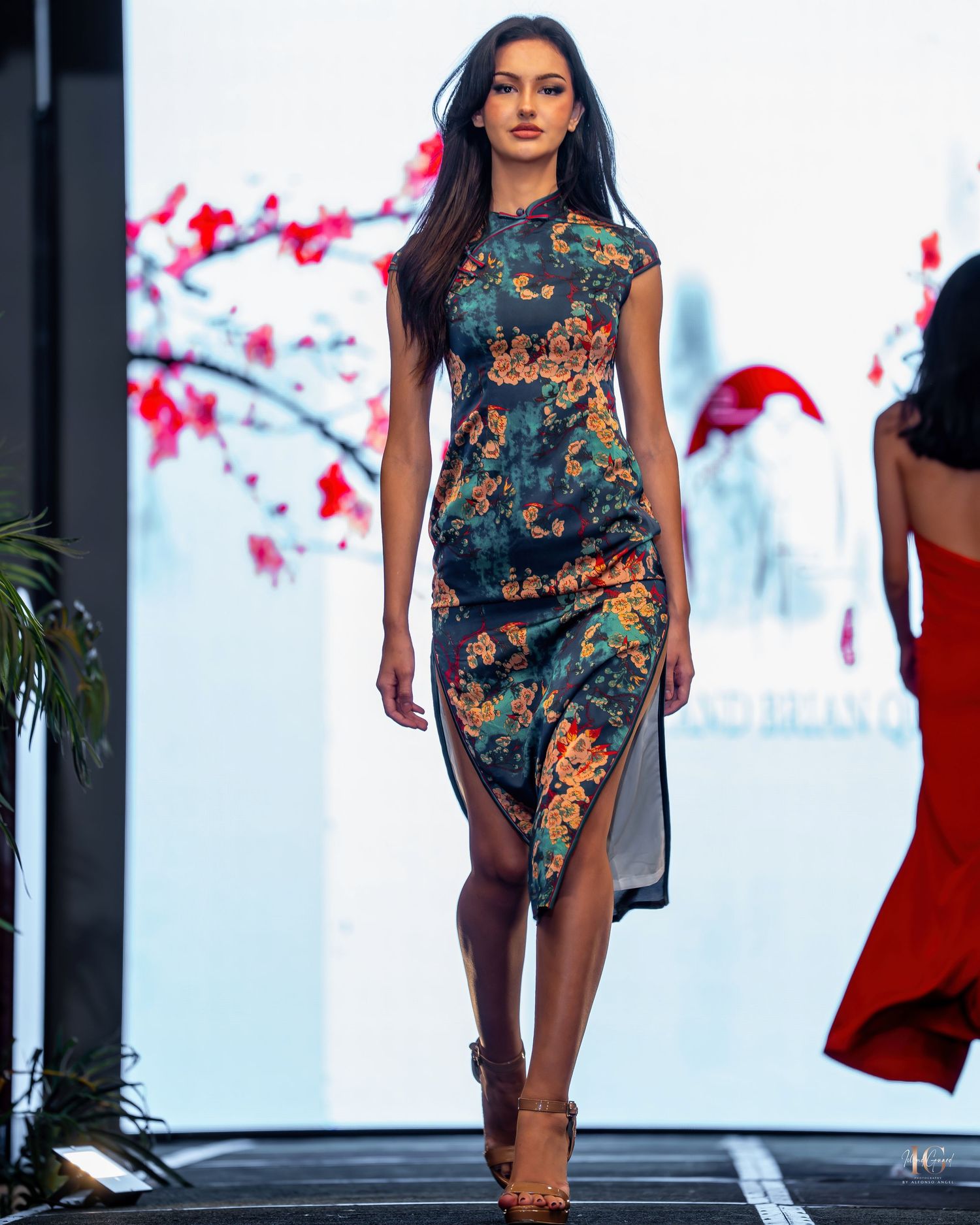 Model walks runway in teal and orange floral print qipao-style dress with side slit against cherry blossom backdrop.