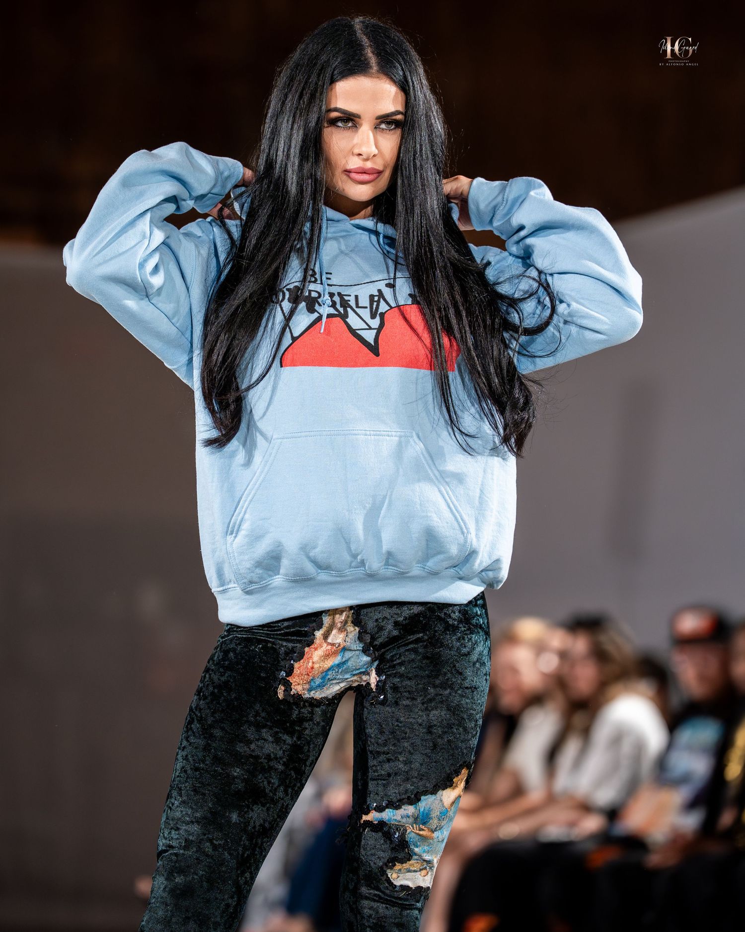 Model walks runway wearing light blue sweatshirt with red stripe and black distressed pants during fashion show.