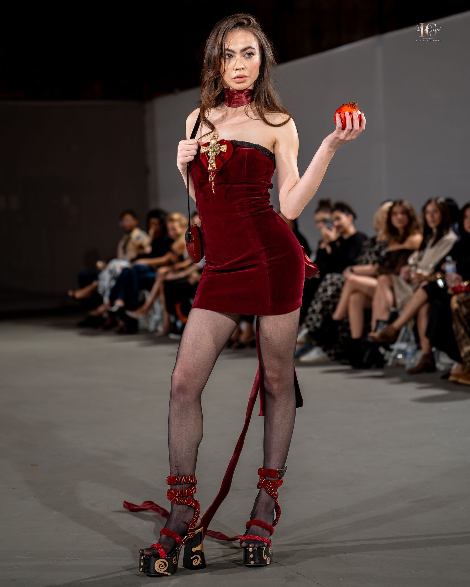 Model walks runway in dark red velvet bodysuit with sheer stockings and strappy heels at fashion show.