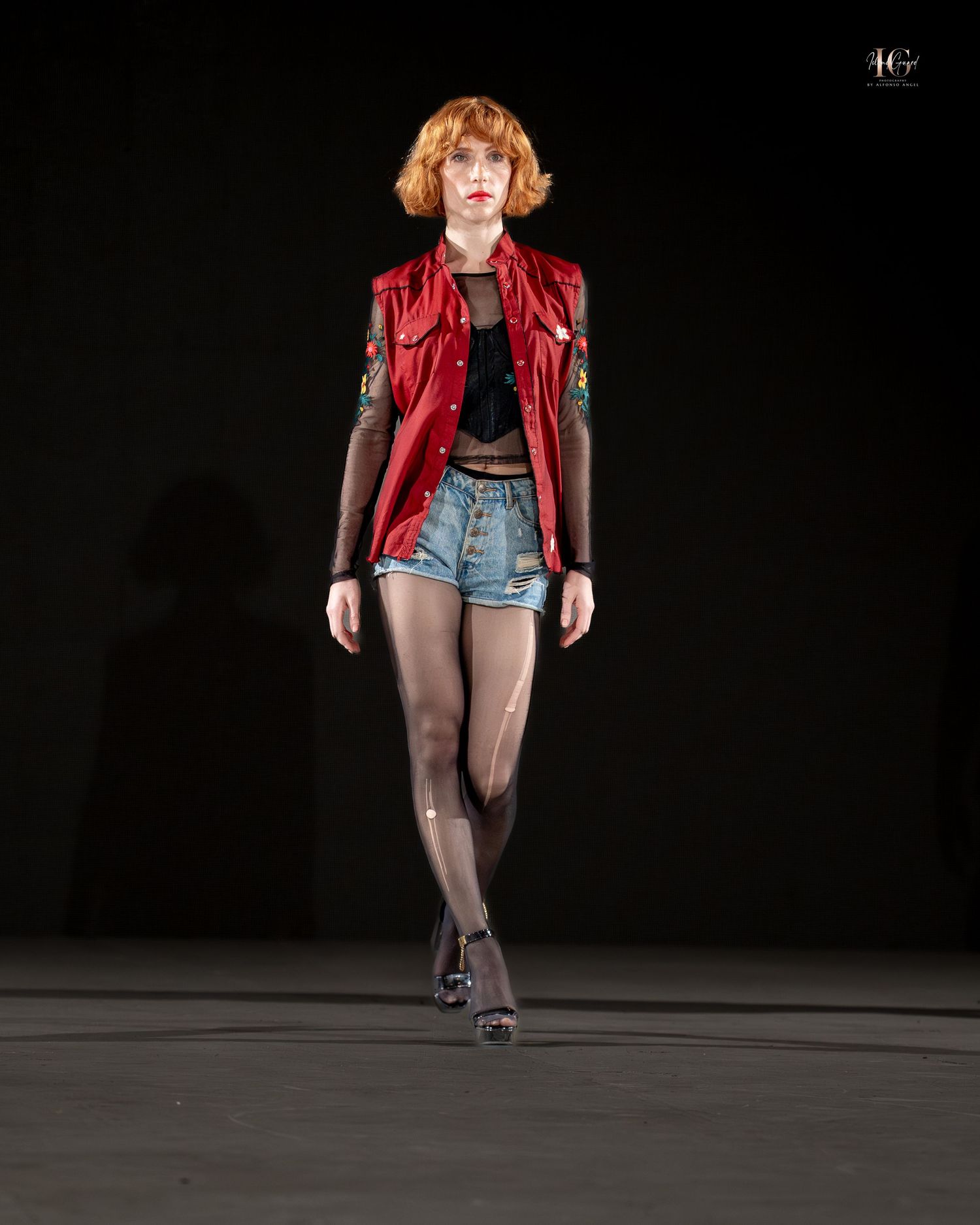 Model walks runway in red leather jacket, denim shorts, and black tights during fashion show presentation.