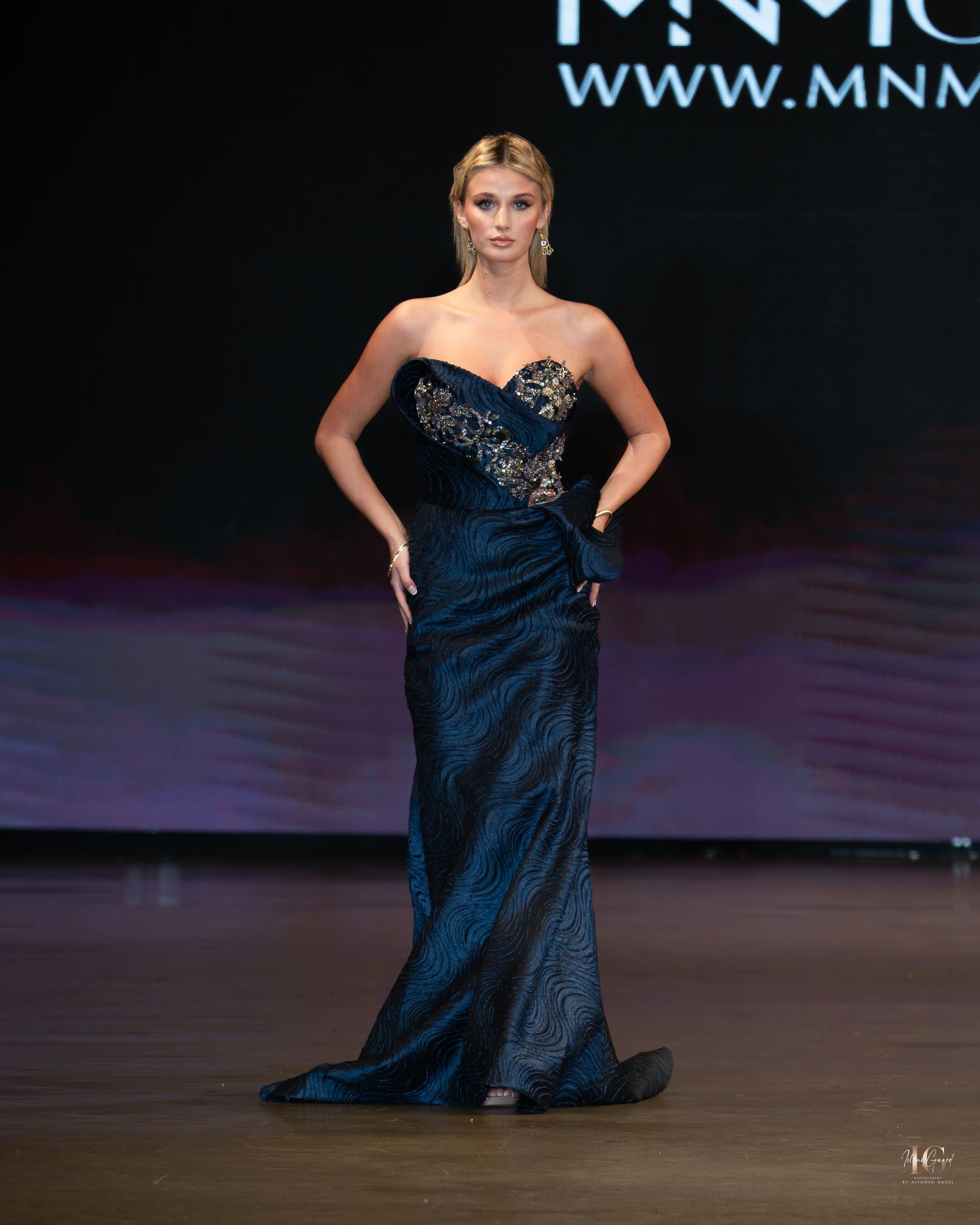 A model walks the runway in a strapless navy blue evening gown with embellished details during a fashion show.