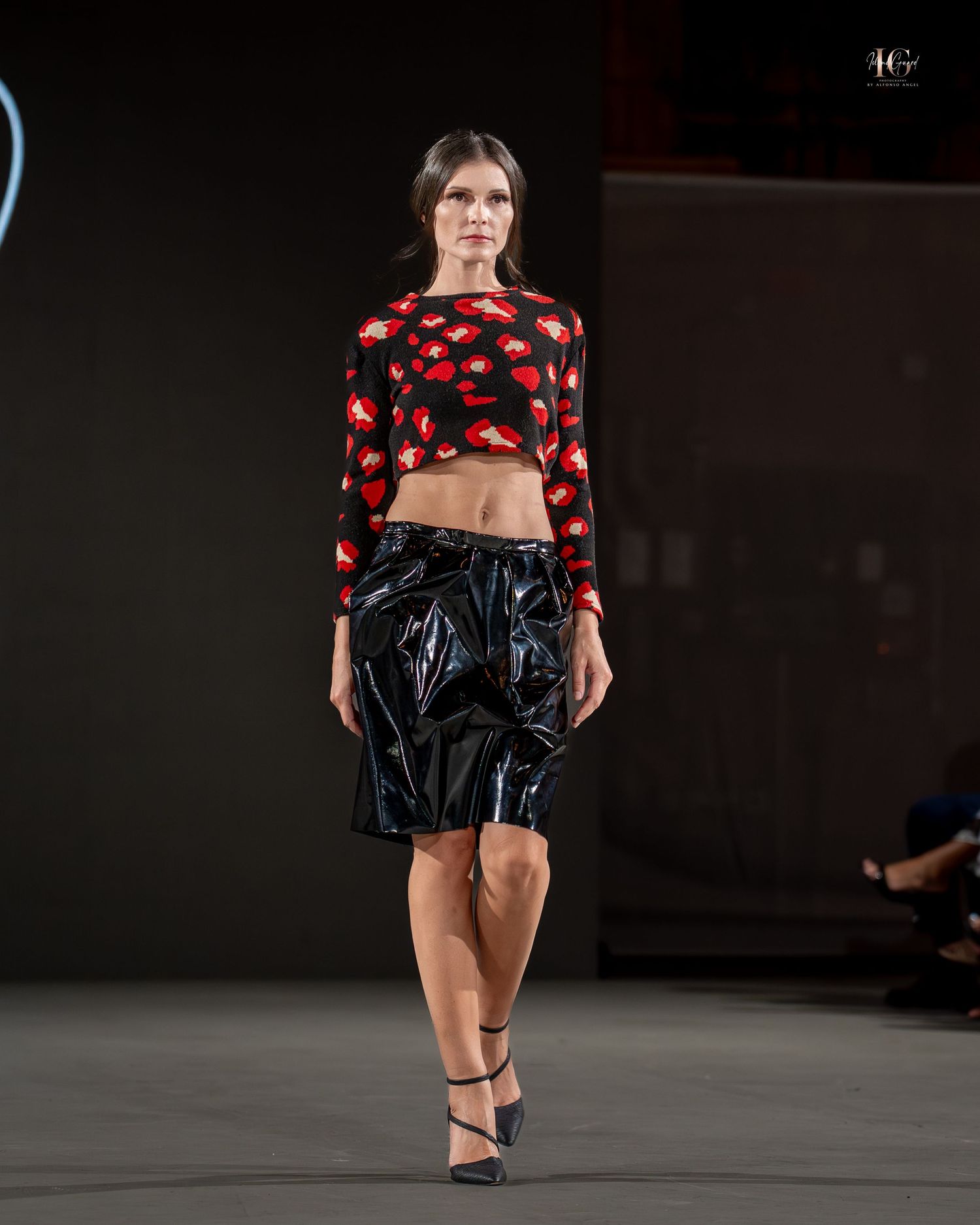 Model walks runway wearing red and black patterned crop top with shiny black knee-length skirt and strappy heels.