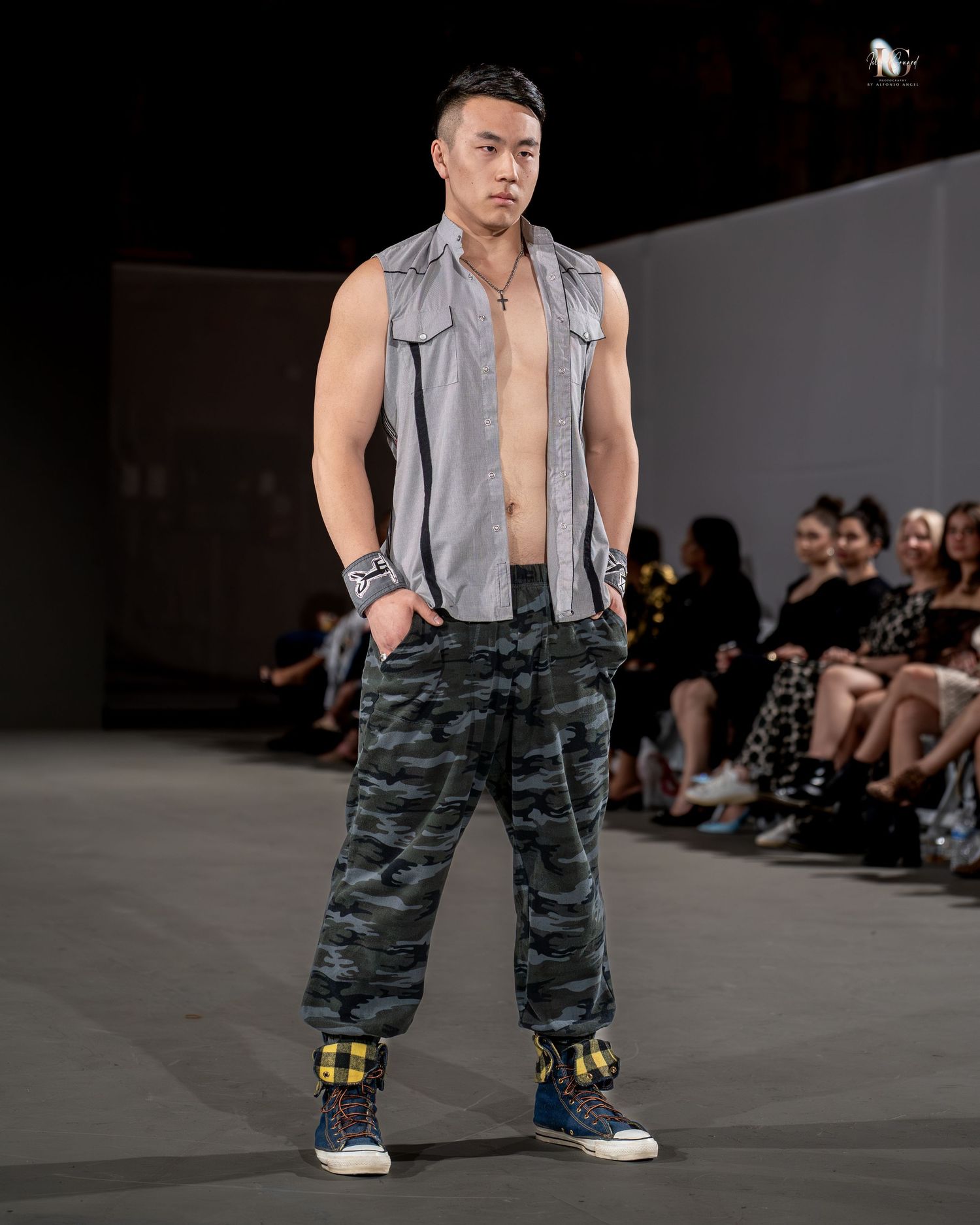 Model wearing an open gray vest and camouflage cargo pants walks runway at fashion show.