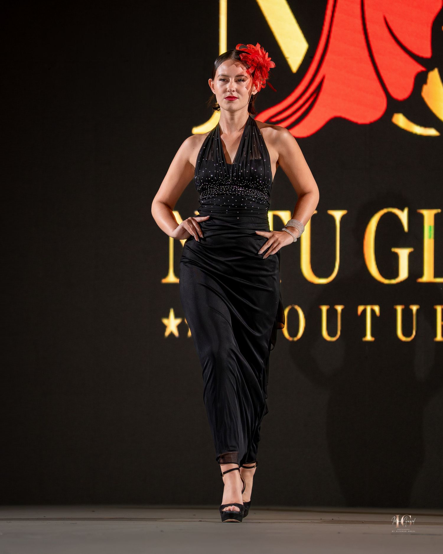A performer in a black halter dress and red flower accessory strikes a pose on a fashion runway with logo backdrop.