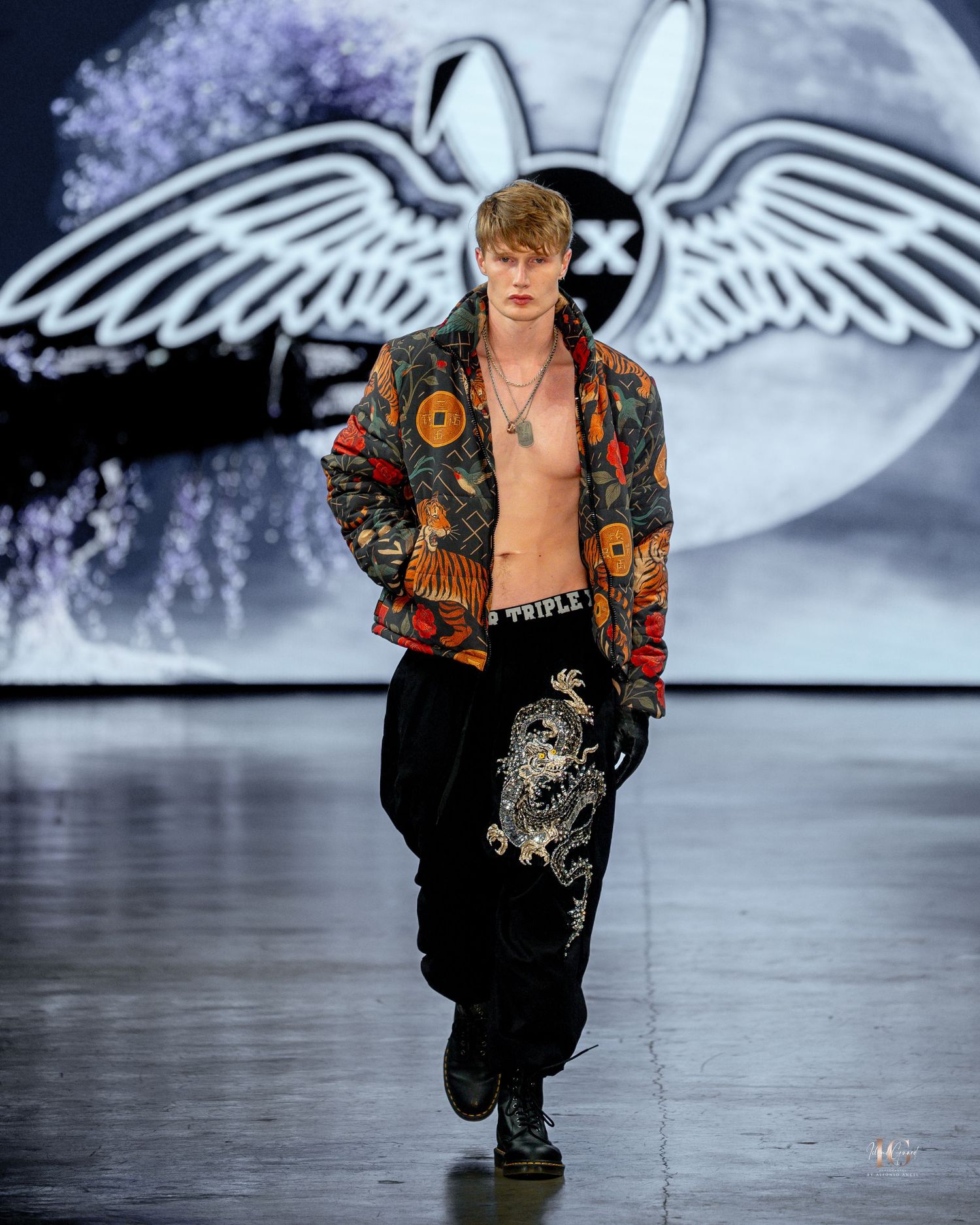 Model walks runway in ornate floral bomber jacket during fashion show with angel wing backdrop.