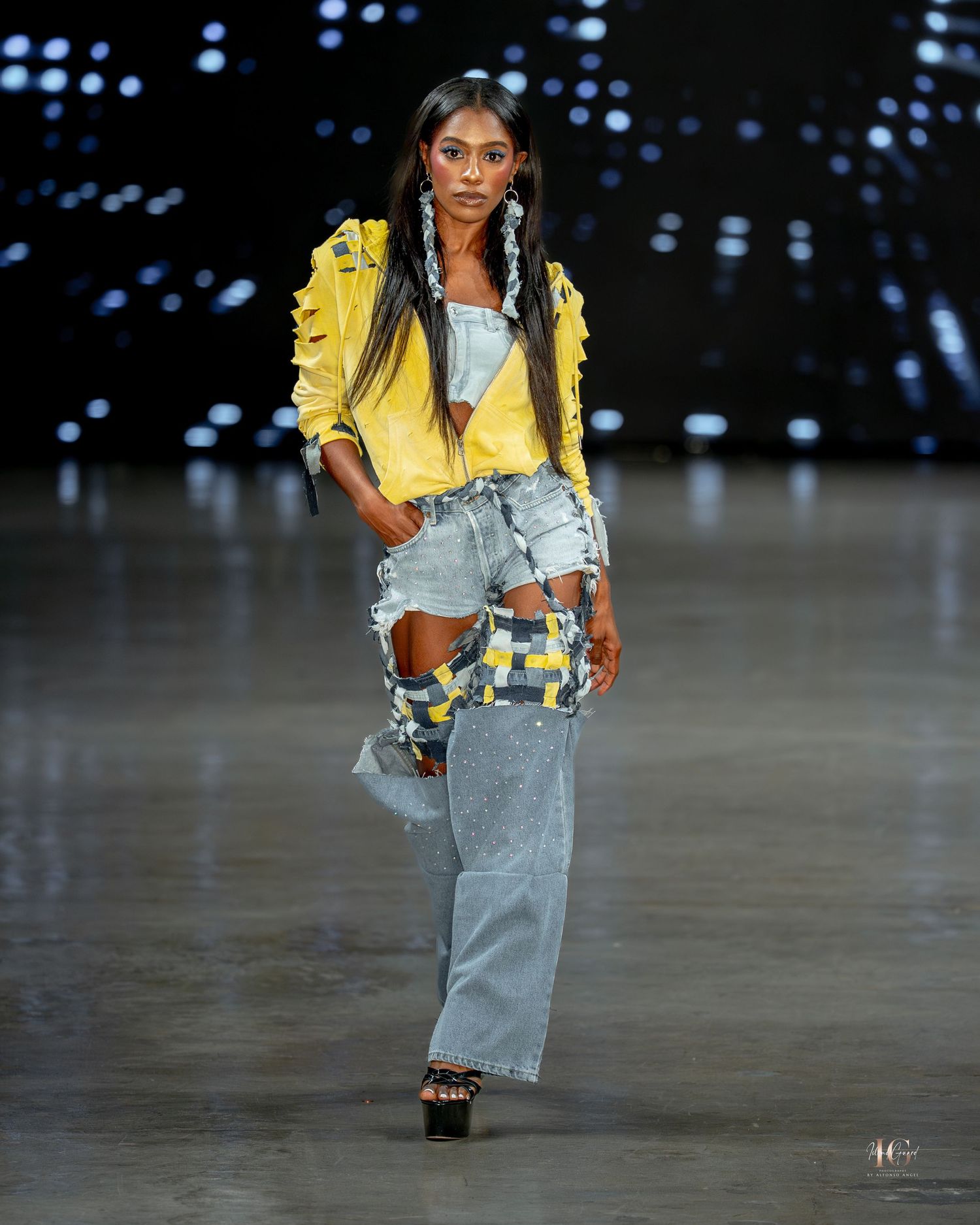 Model walks runway in yellow jacket and distressed wide-leg jeans during fashion show with dark lighting effects.