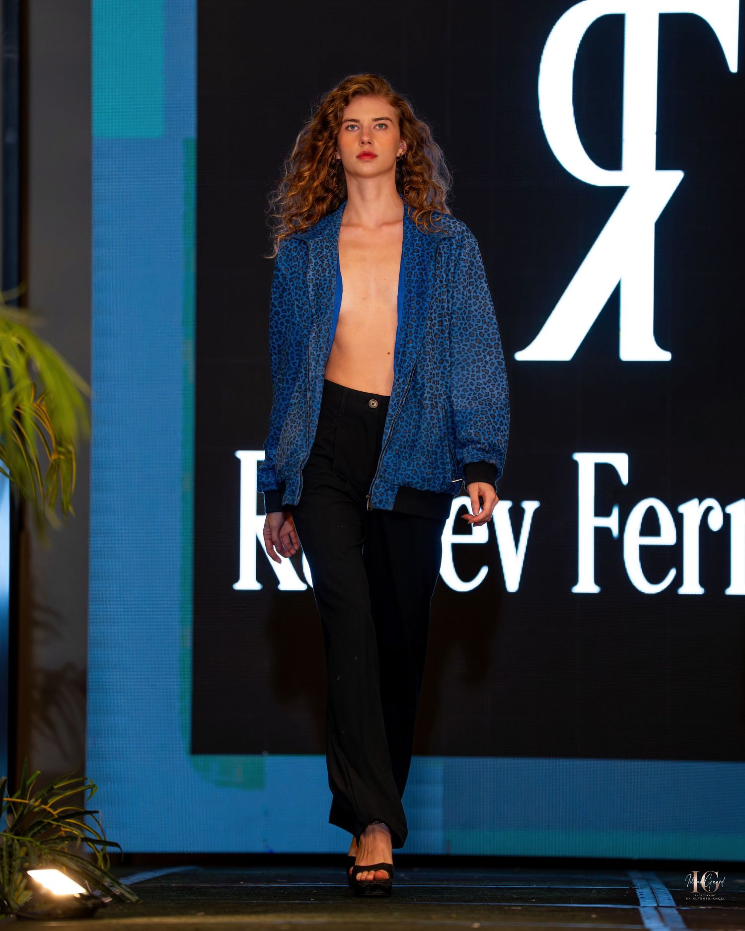 Model wearing a blue blazer walks down a dark runway with illuminated white stripes and 'R' lettering in the background.