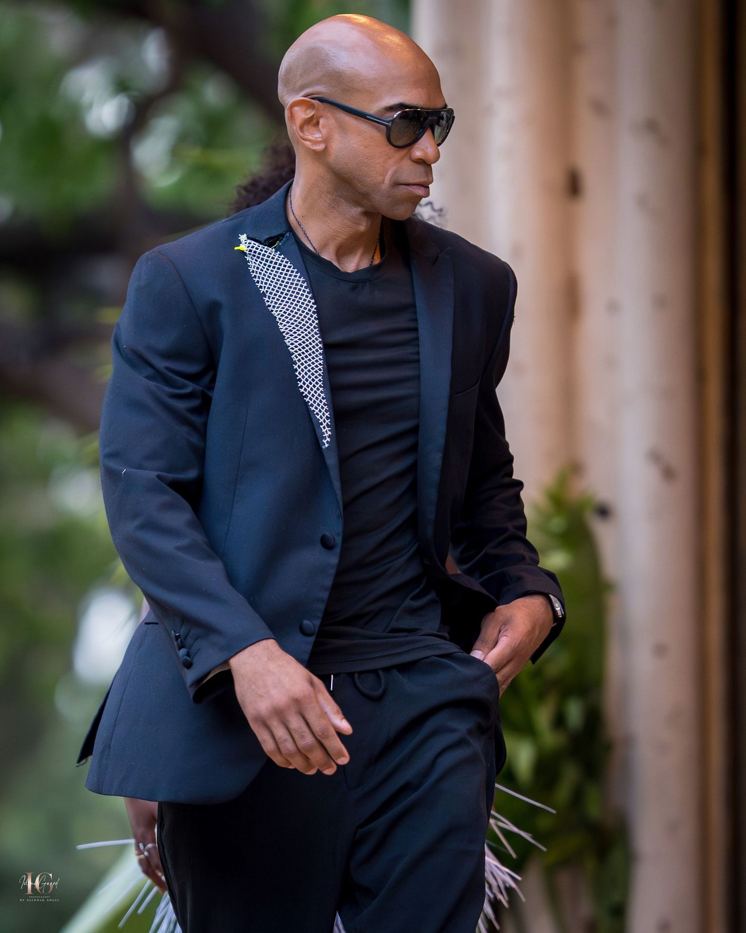 A person wearing a navy blazer and dark sunglasses walks outdoors while carrying shopping bags.