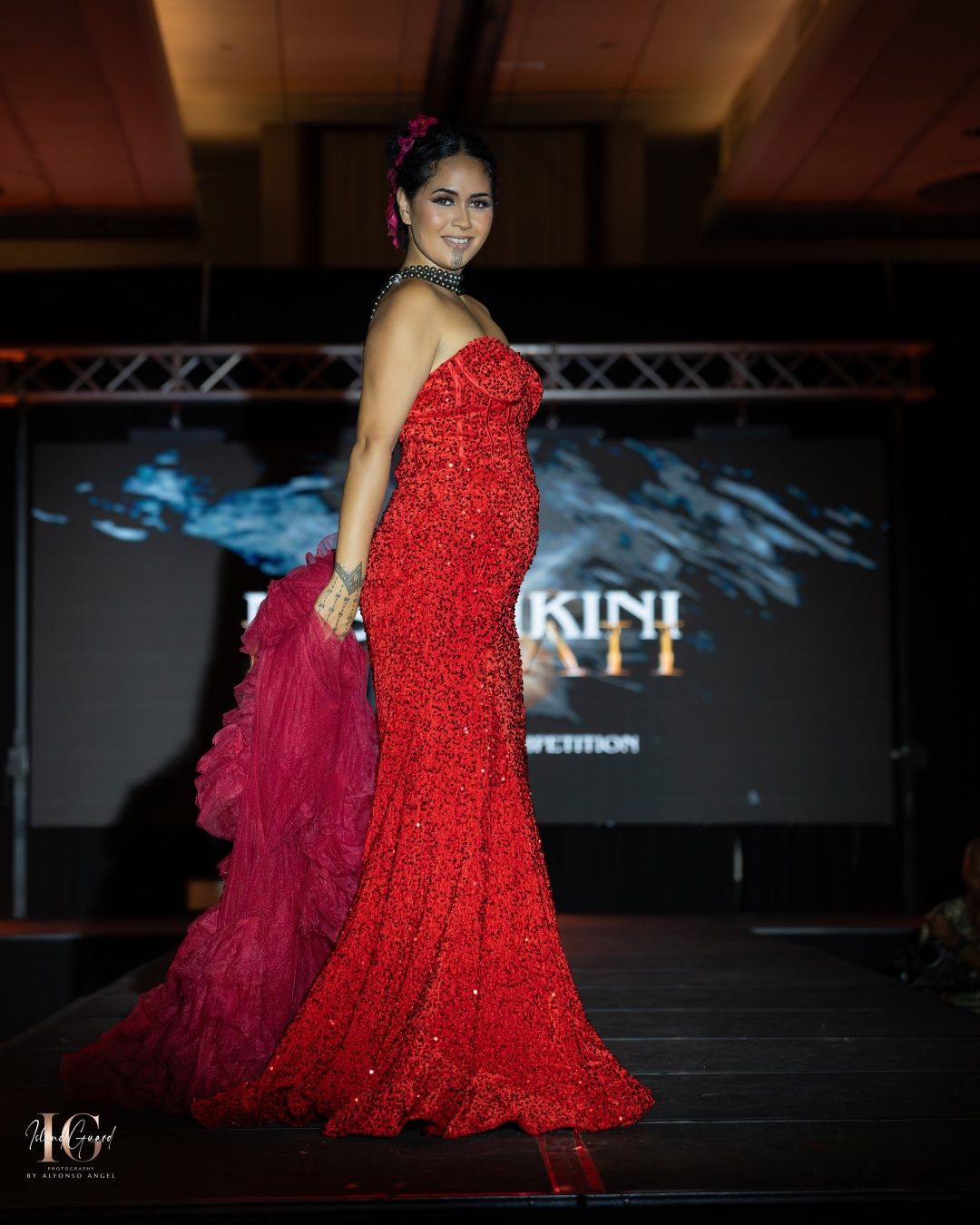 Model walks the runway in a stunning red sequined evening gown with burgundy accessory at a fashion show.