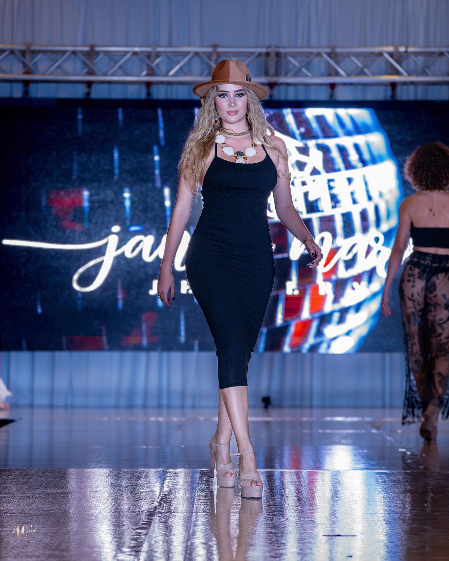 Model showcases sleek black midi dress and cowboy hat on illuminated runway.