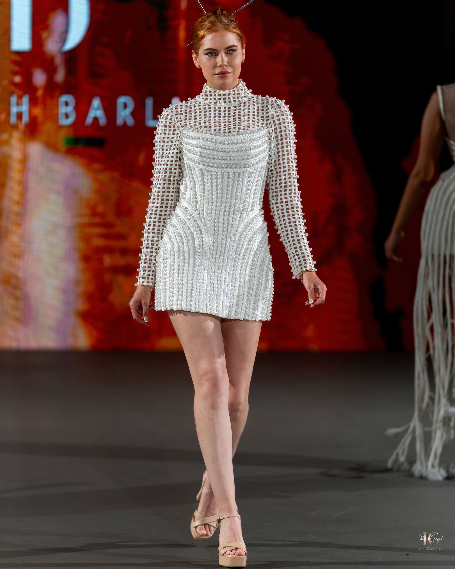 Model walks runway in white crochet mini dress with fringe details at fashion show against red backdrop.