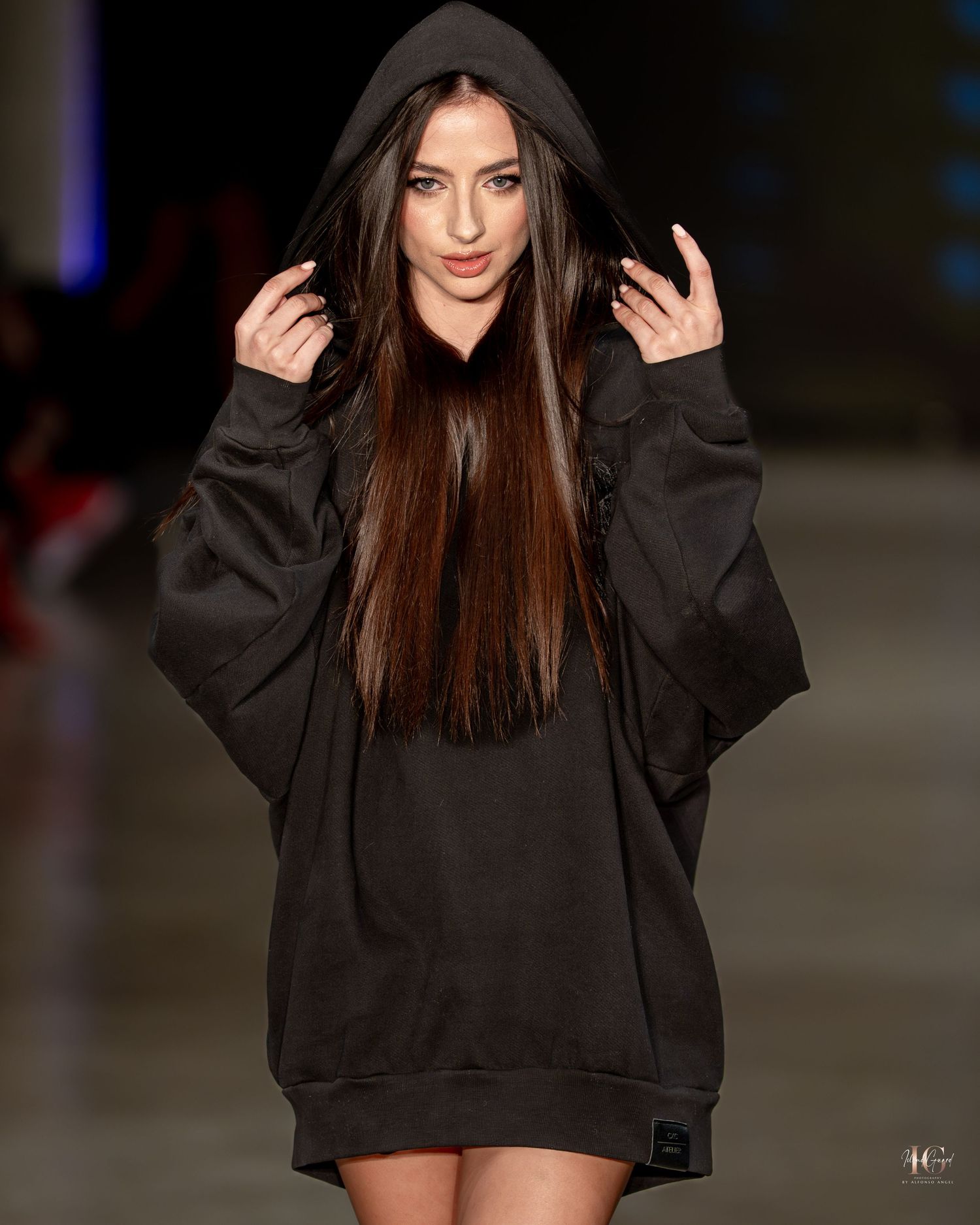 Model wearing an oversized black hoodie dress with long straight dark hair walks the runway during fashion show.