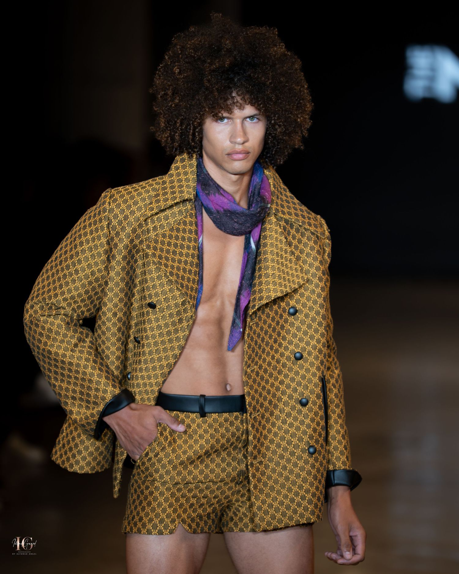Fashion model walks runway wearing yellow patterned cropped jacket with purple scarf at fashion show.