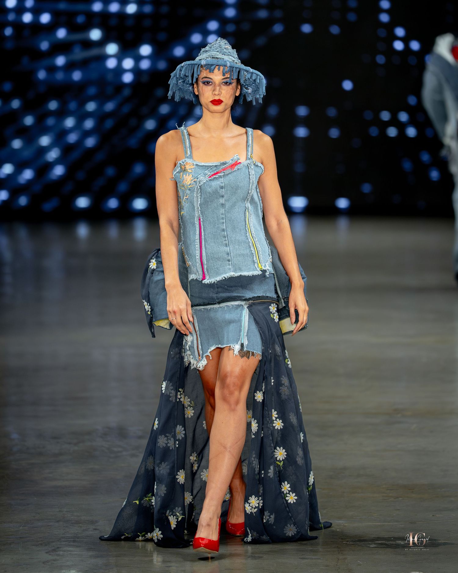 Fashion model walks runway in distressed denim outfit with flowing floral skirt and red heels during fashion show.