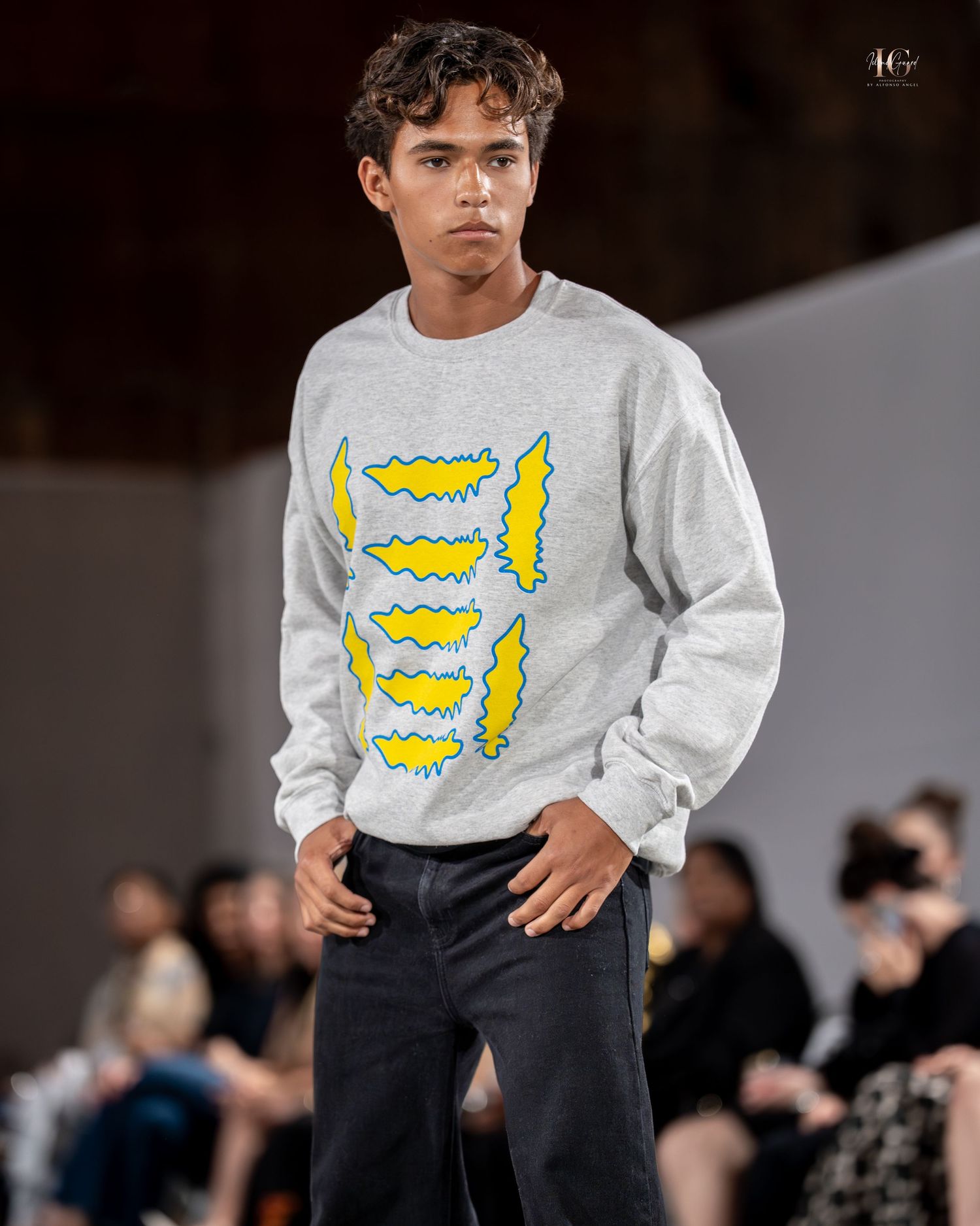 Model walks runway wearing light gray sweatshirt with yellow graphic design and dark pants during fashion show.