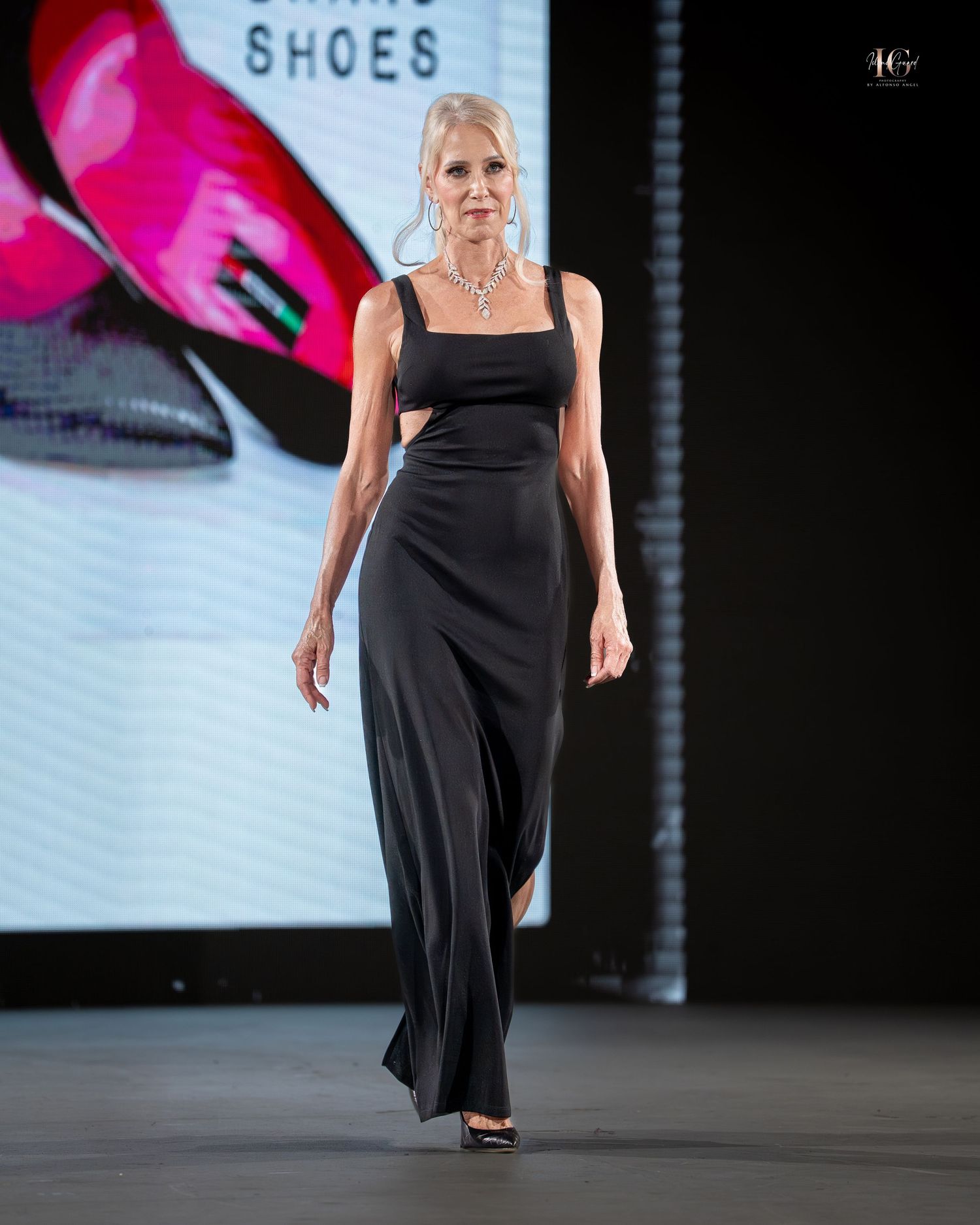 Model walks runway in black evening wear during fashion show presentation with pink lighting accents.