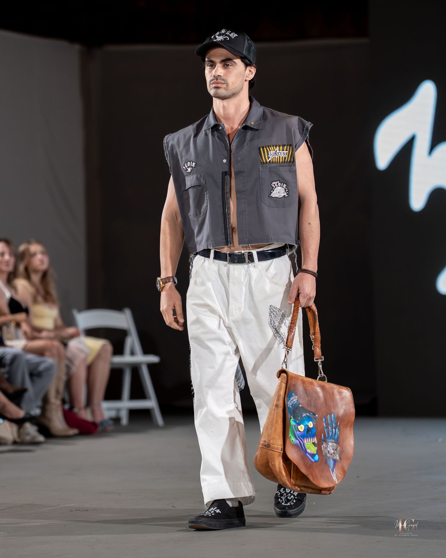 Model wearing white pants, grey vest, and baseball cap walks runway with brown leather messenger bag at fashion show.