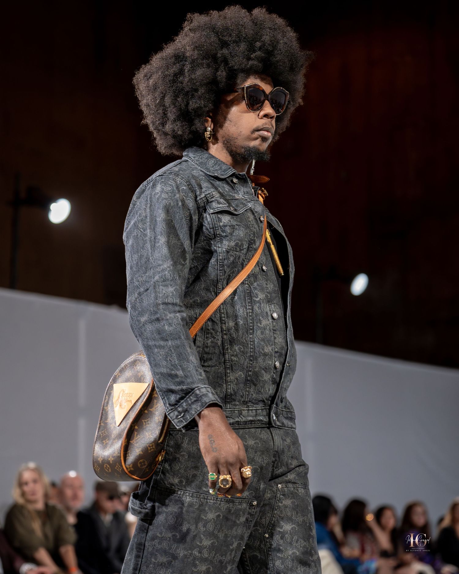 Model walks the runway wearing a gray denim ensemble with sunglasses and a brown leather bag at a fashion show.