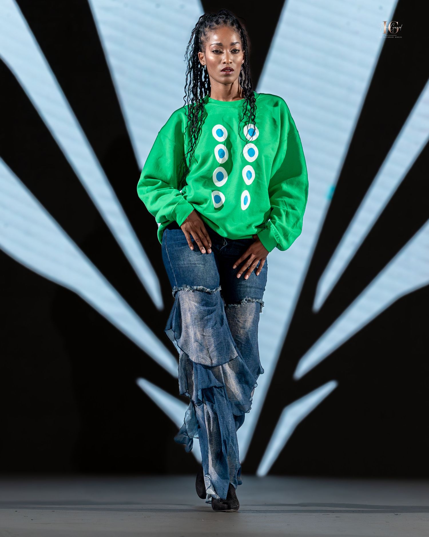 Model walks runway wearing bright green sweatshirt and distressed baggy jeans against black and white striped backdrop.