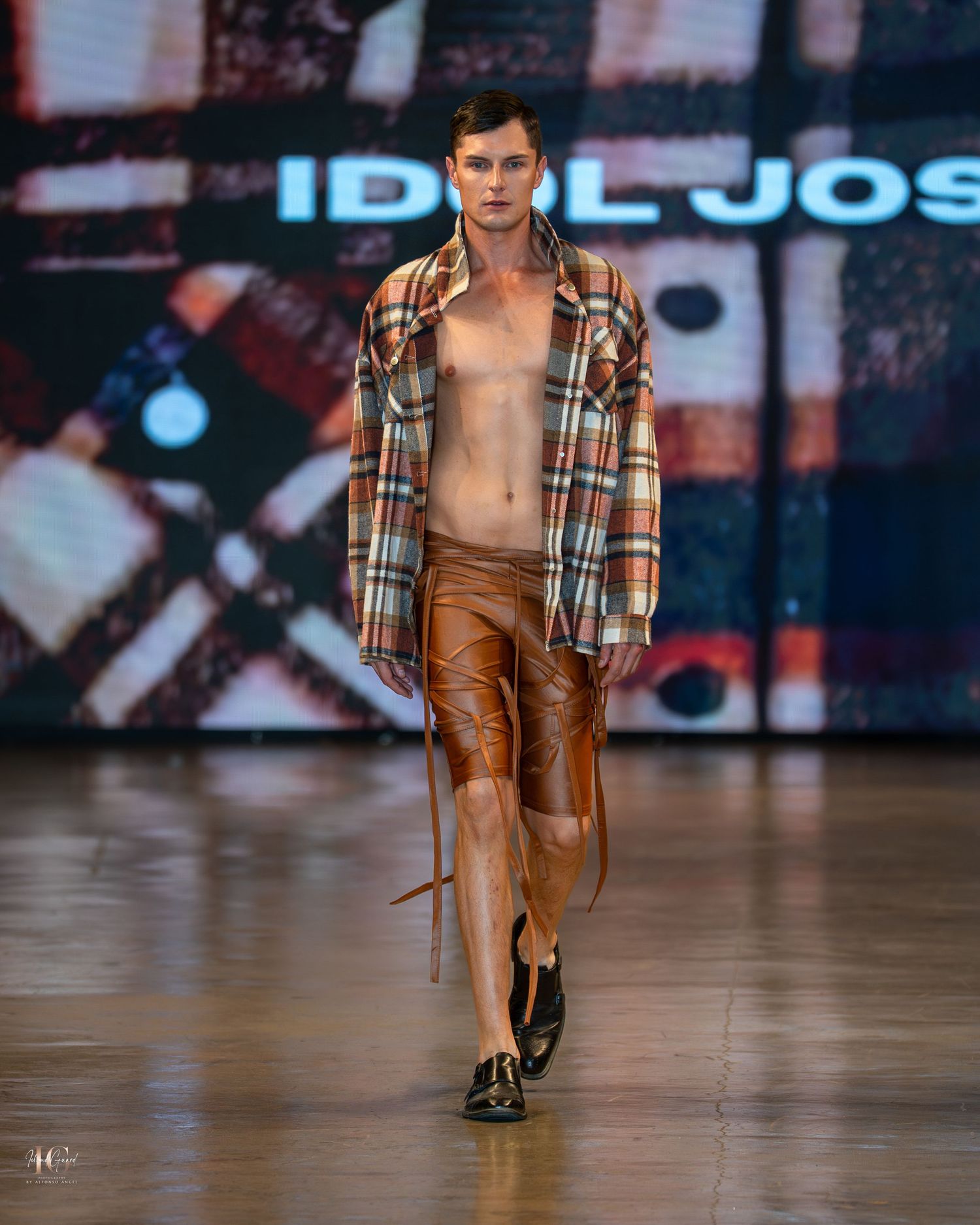 Model walks runway in brown leather pants and plaid button-down shirt during fashion show.