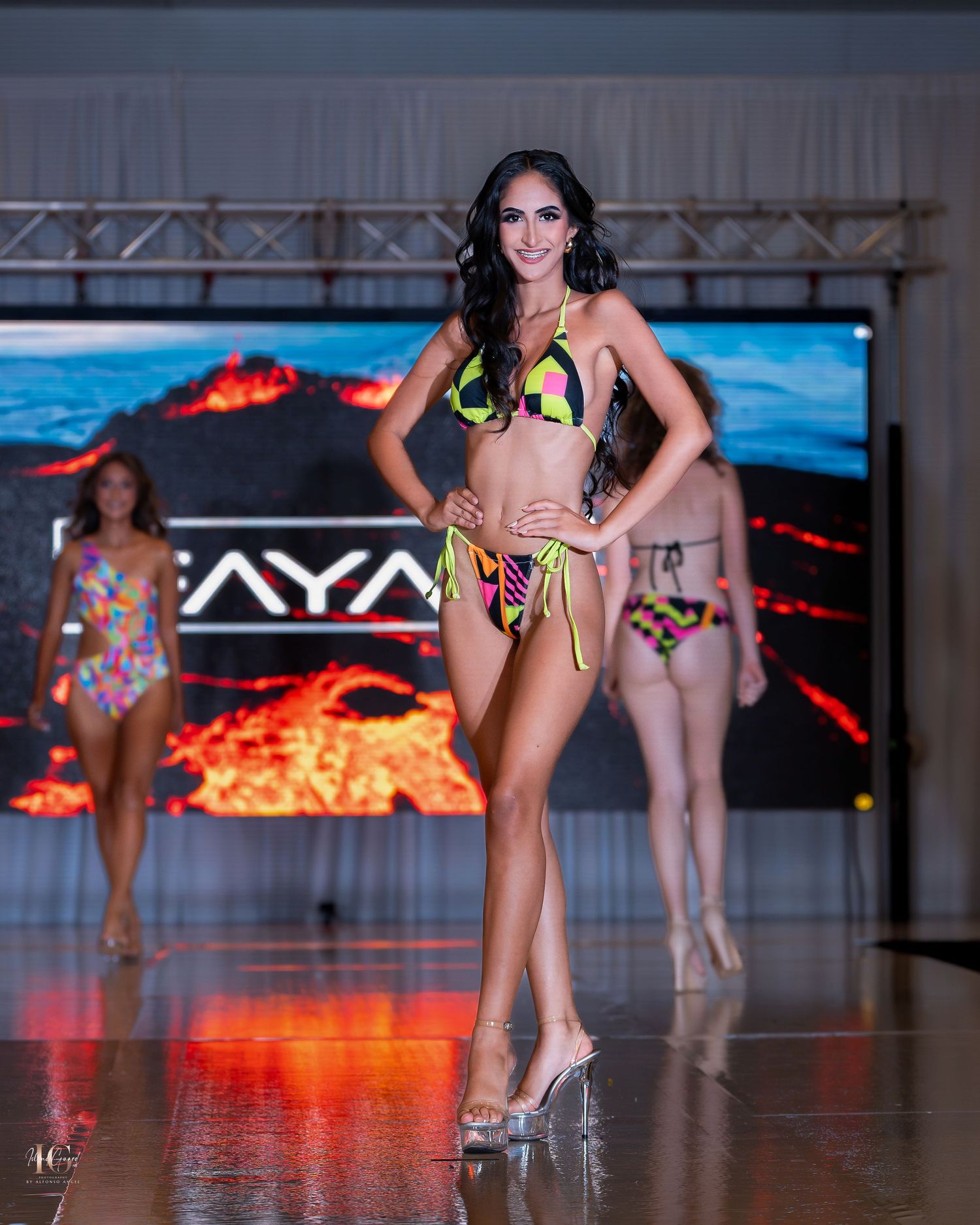 Model walks the runway in a neon yellow bikini with tropical print during a swimwear fashion show with volcanic backdrop.