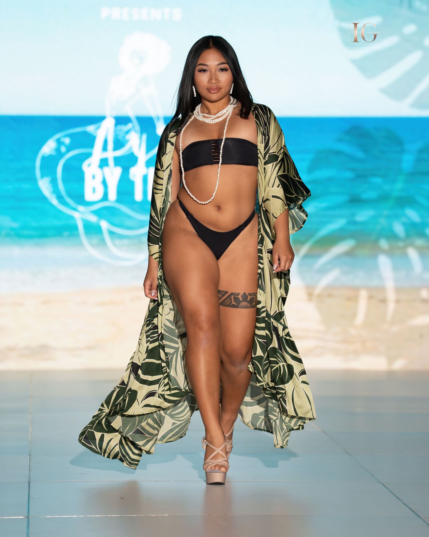Model in black swimwear and tropical print cover-up poses on beach-themed runway during fashion show.