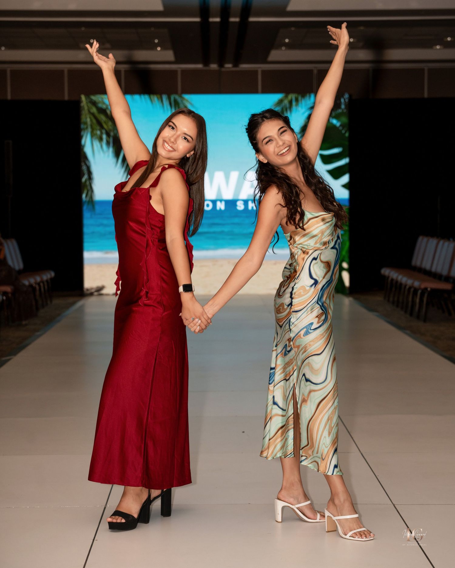 Two models walk together on a fashion runway, one wearing a burgundy dress and another in a patterned beige outfit.