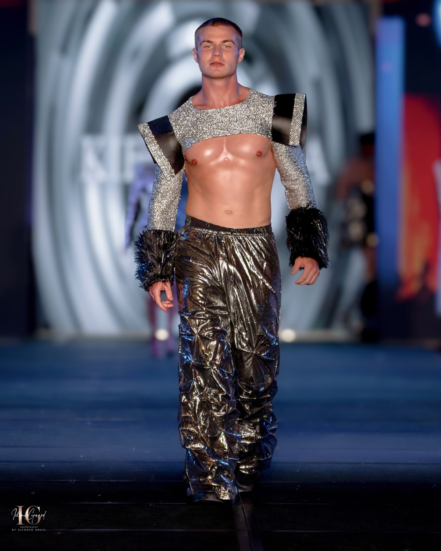 Model wearing metallic silver crop top and shiny brown pants on fashion runway with circular backdrop.