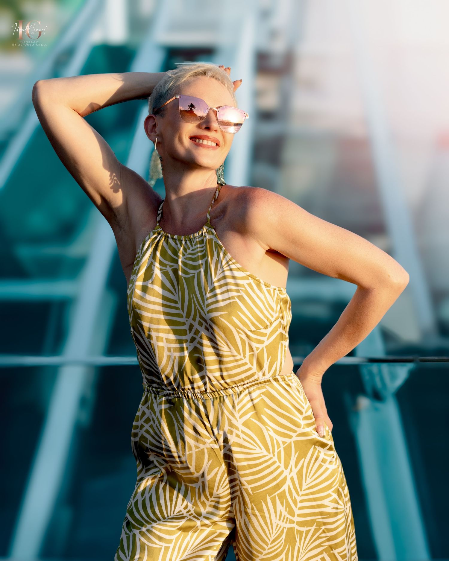 Someone in a summery yellow and white palm print halter jumpsuit poses against a turquoise background in bright sunlight.