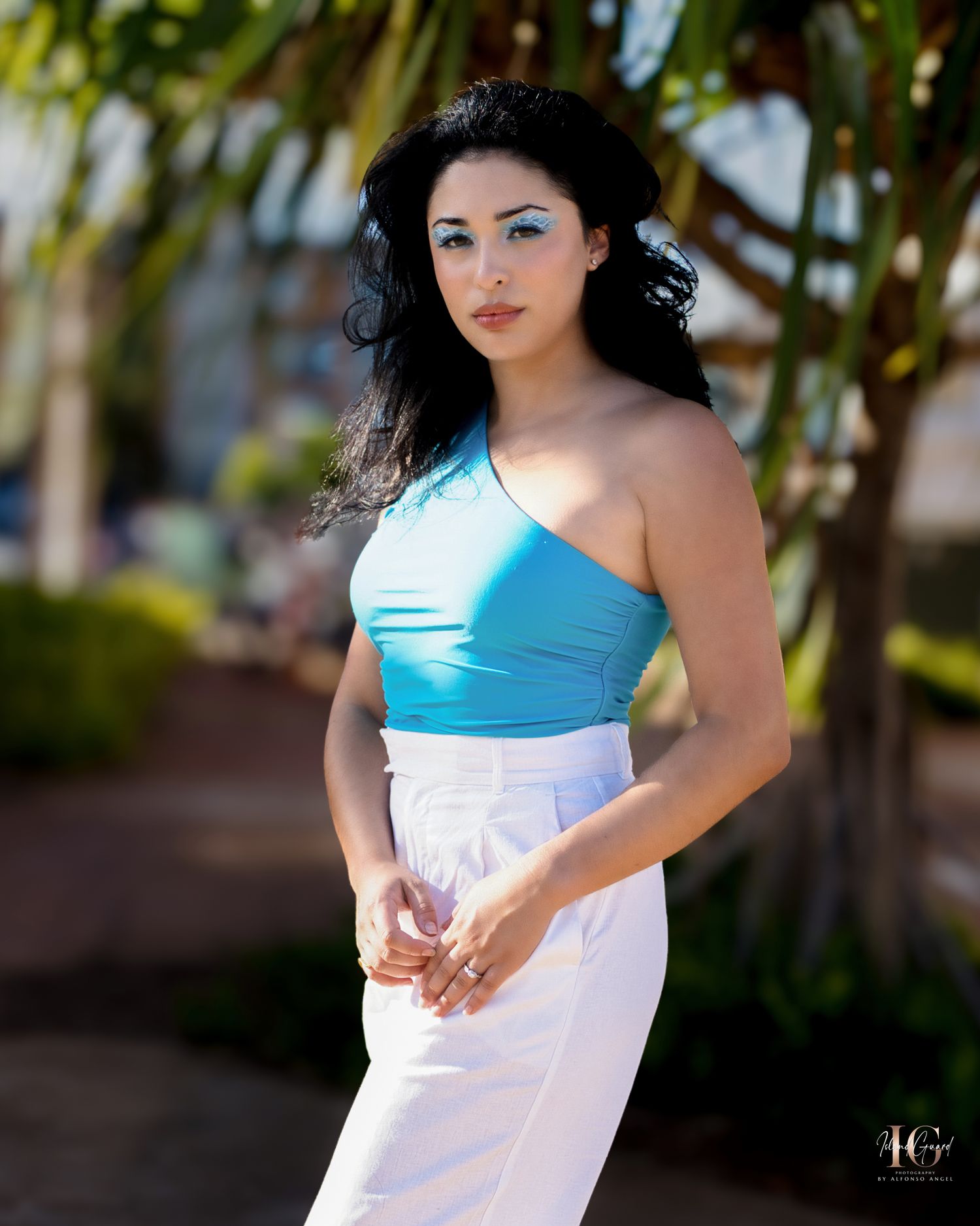 A person in a light blue one-shoulder top and white skirt poses outdoors near palm trees in bright sunlight.