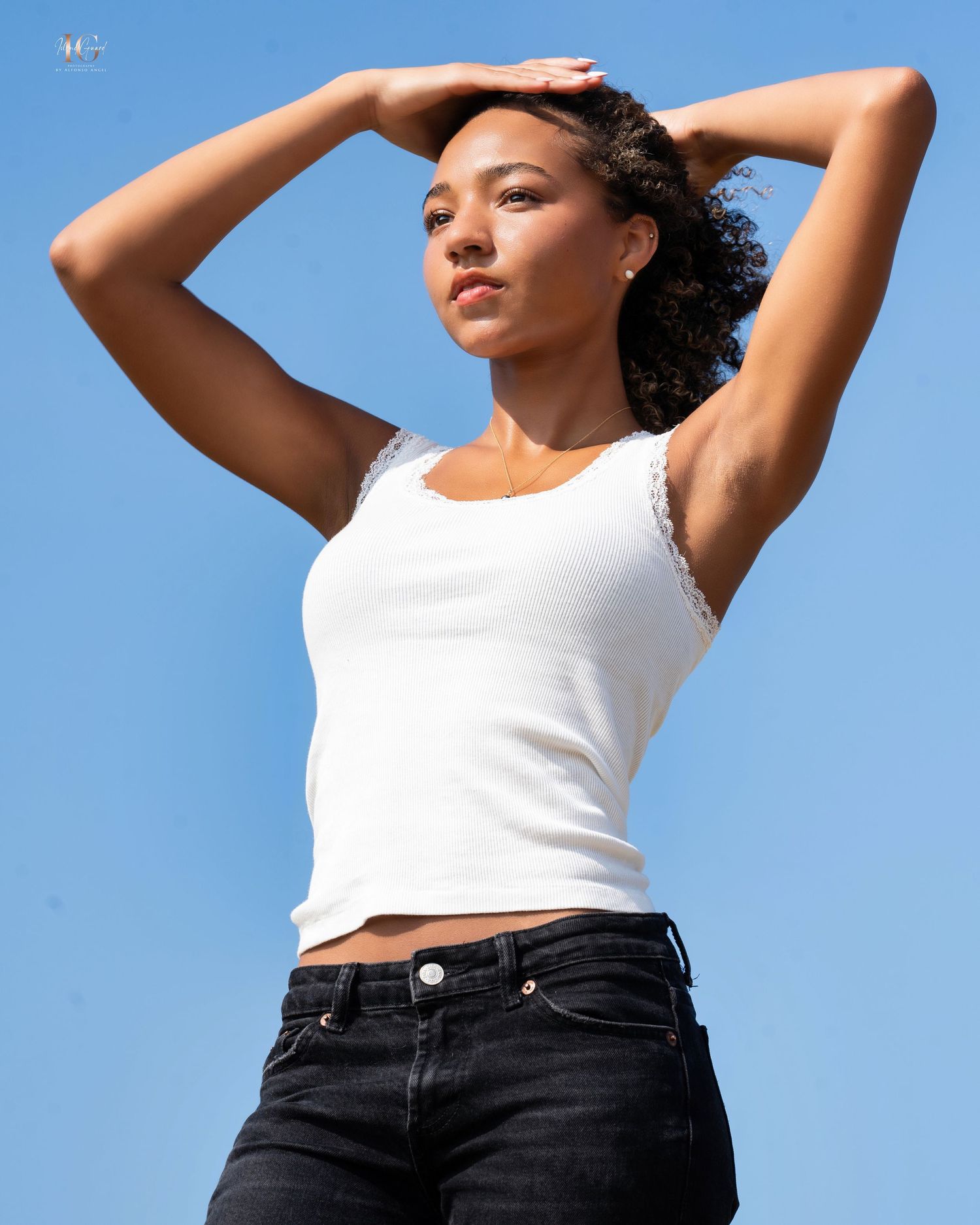 A person in a white tank top and dark jeans poses expressively against a blue sky background in a casual photoshoot.
