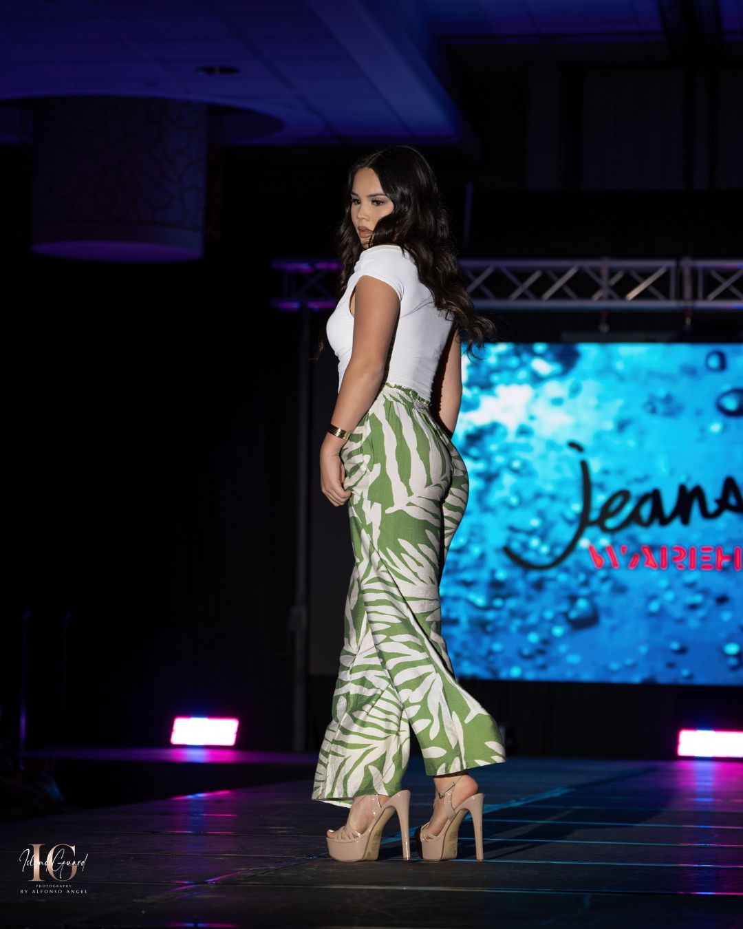 Model showcases tropical print dress on runway with blue digital backdrop.