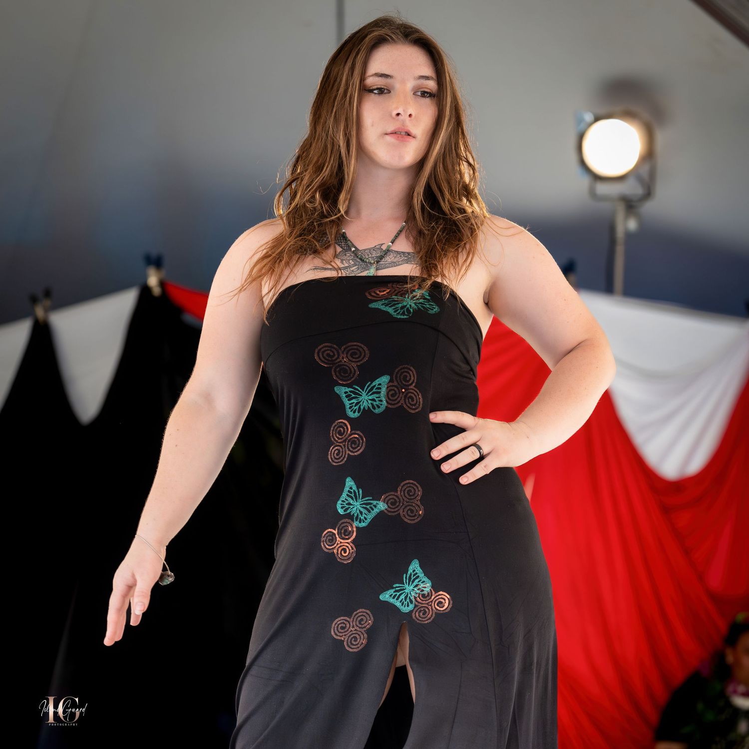Model wearing black evening dress with butterfly embellishments on runway against red backdrop.